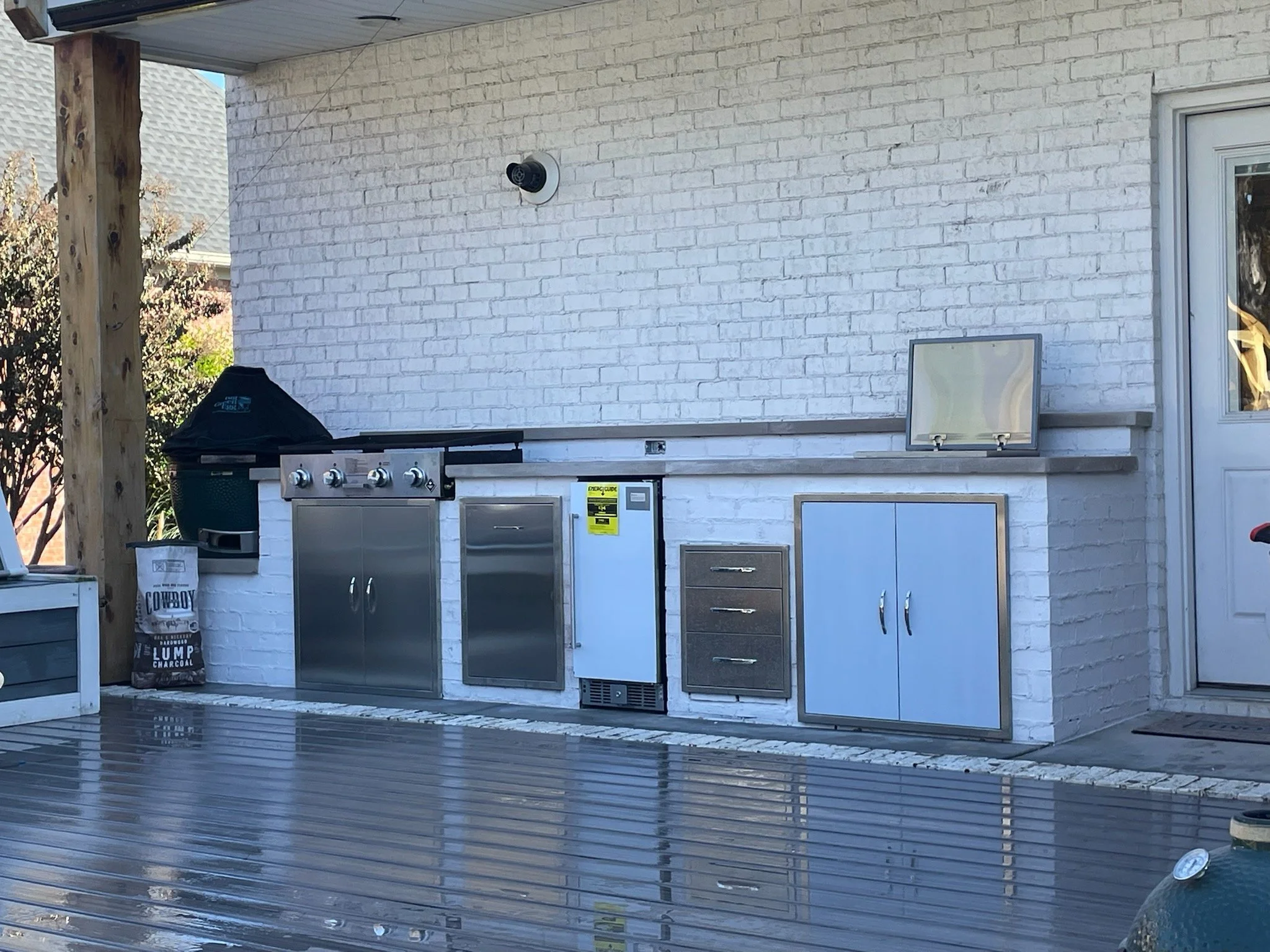 Outdoor patio area with a built-in grill, cooking utensils, storage cabinets, a small refrigerator, and a speaker, set against a white brick wall with a door to the right.