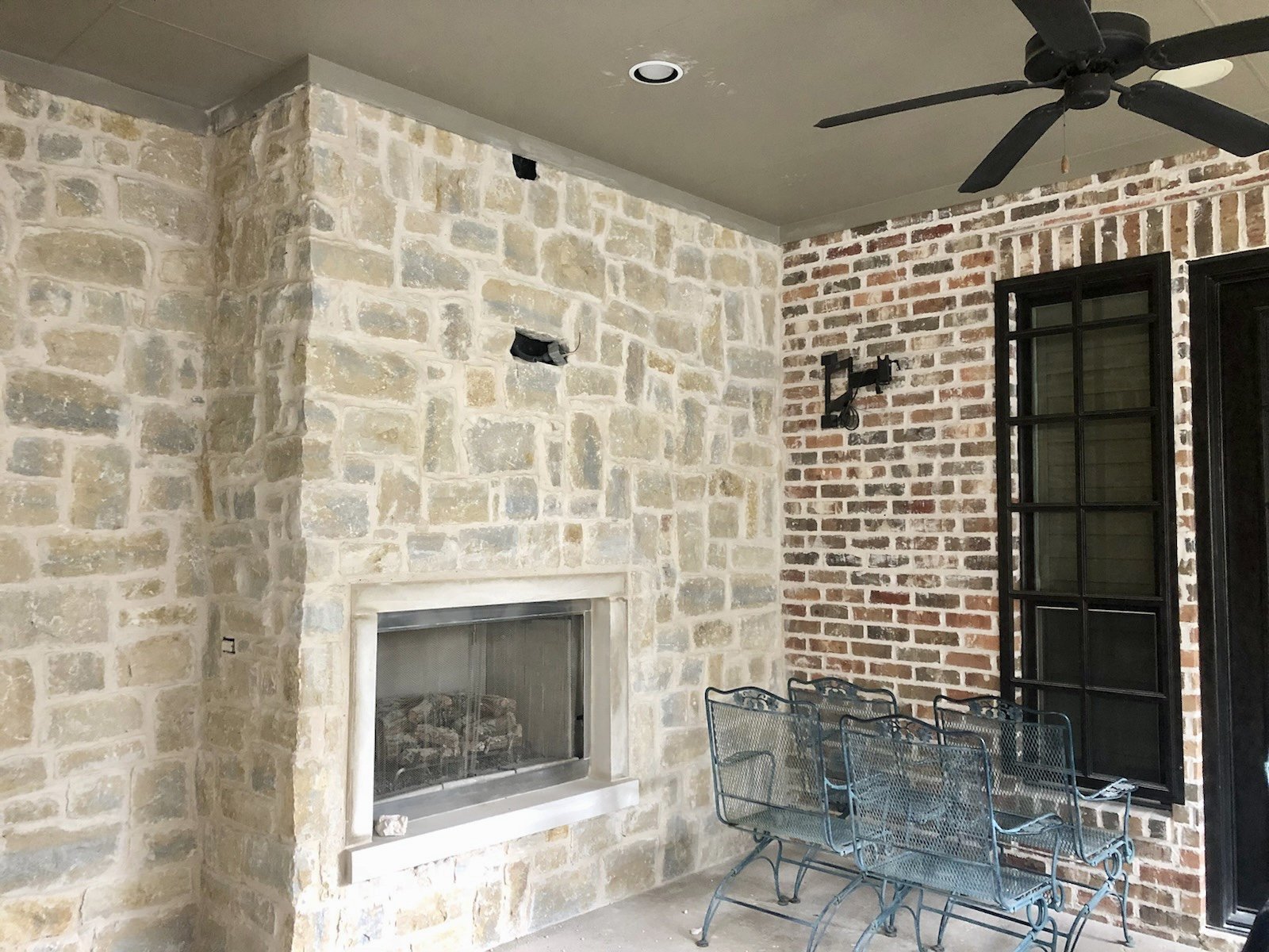Outdoor patio with stone fireplace, wrought iron chairs, ceiling fan, and brick wall.