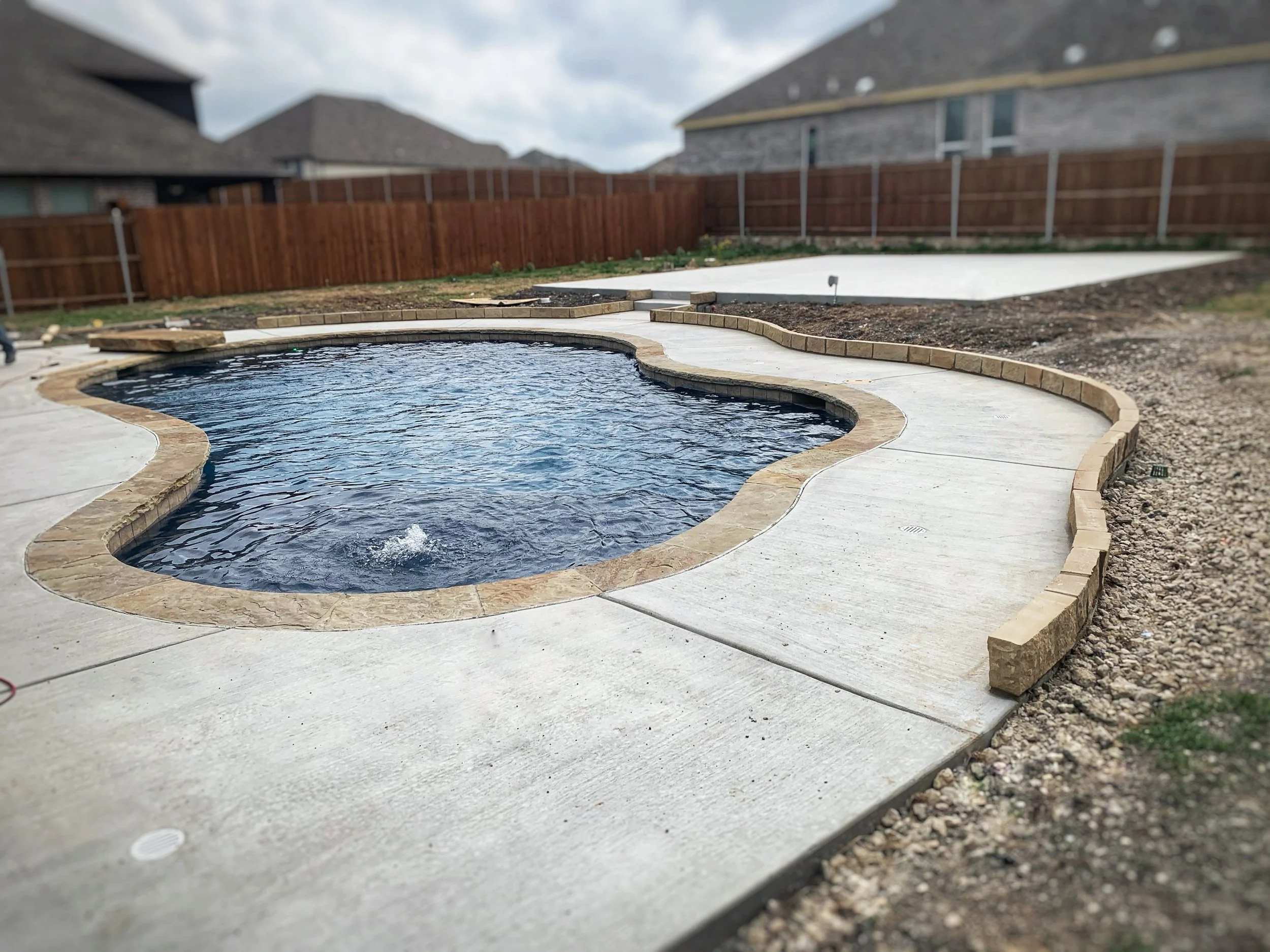 Newly constructed backyard swimming pool with curved concrete deck and stone border, surrounded by a wooden fence. There are neighboring houses visible in the background.