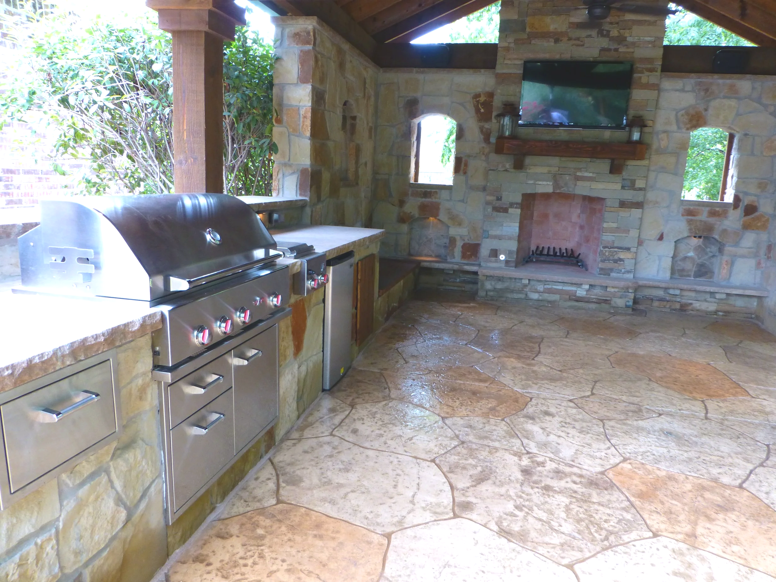 Outdoor kitchen area with a stainless steel grill, stone countertops, and a stone fireplace with a mounted TV above it, surrounded by windows and greenery.