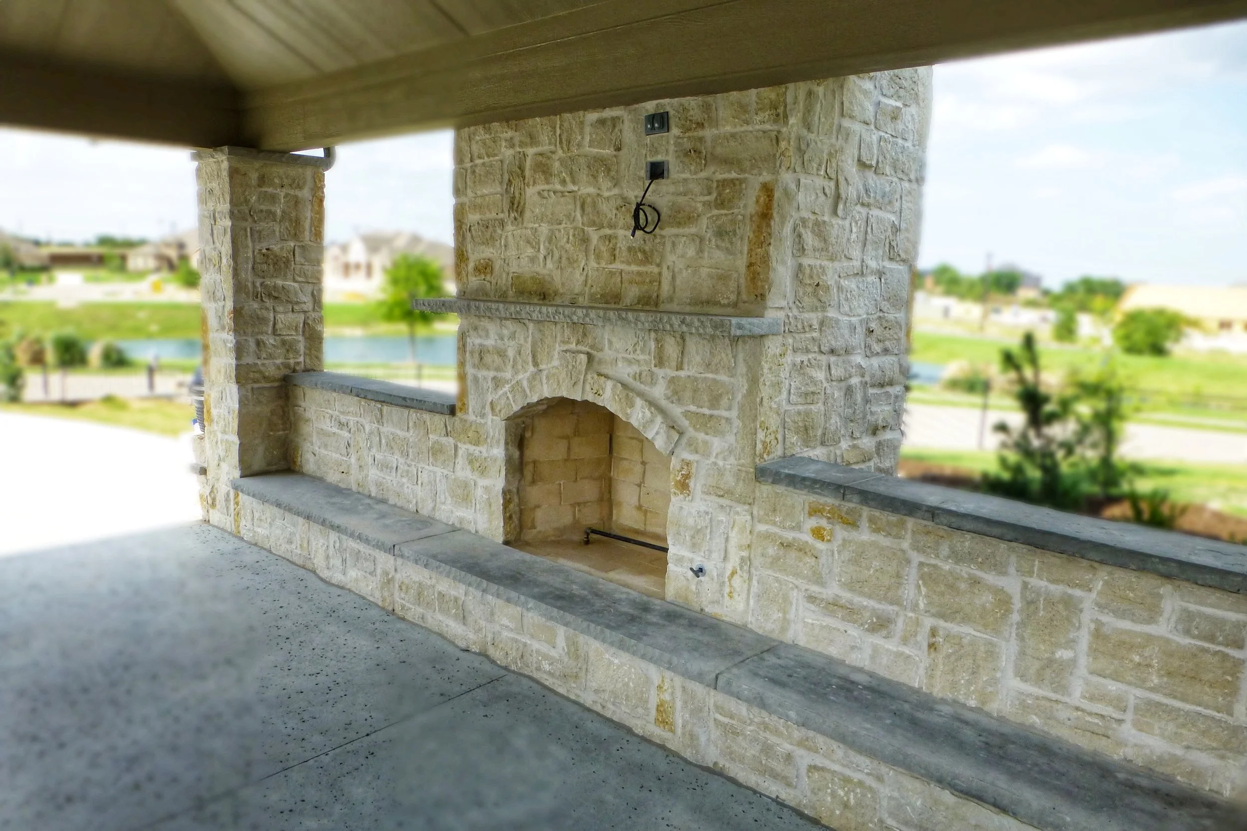 Exterior covered porch area with stone fireplace and ledge, overlooking a scenic lake with houses and trees in the background.