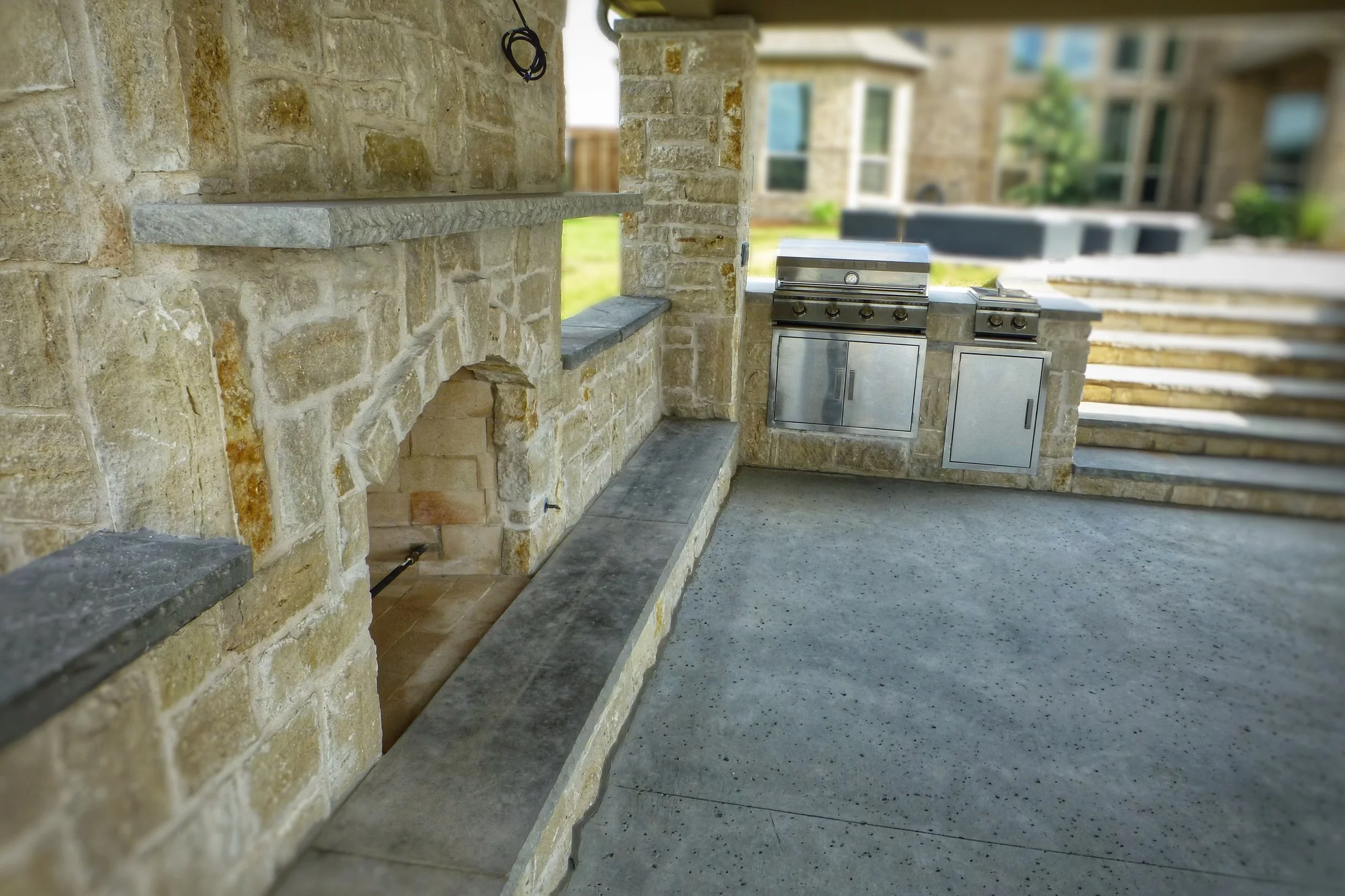 Outdoor kitchen with stonework, a built-in grill, and stairs leading up to a yard with residential buildings in the background.