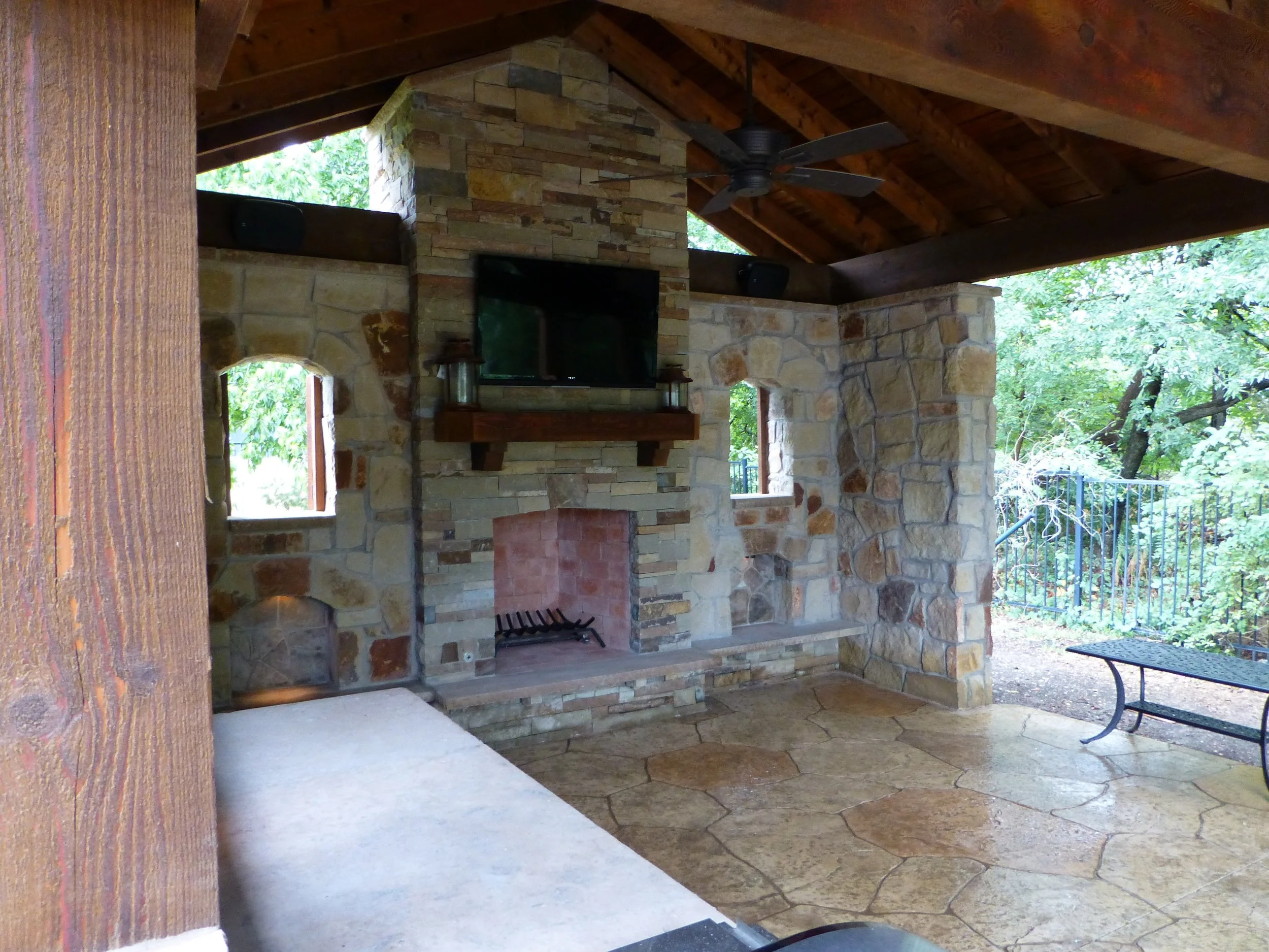 Outdoor covered patio with stone fireplace, flat-screen TV, ceiling fan, and nearby bench, surrounded by trees.