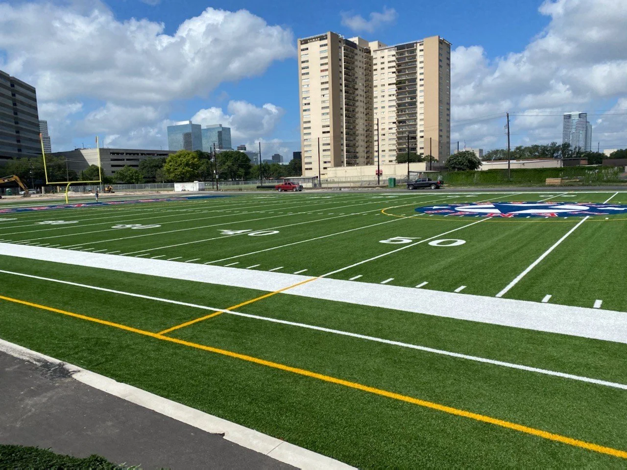 A football field with artificial turf featuring yard line markers and a painted logo at the 50-yard line. The field is situated in an urban area with tall buildings in the background under a partly cloudy sky.