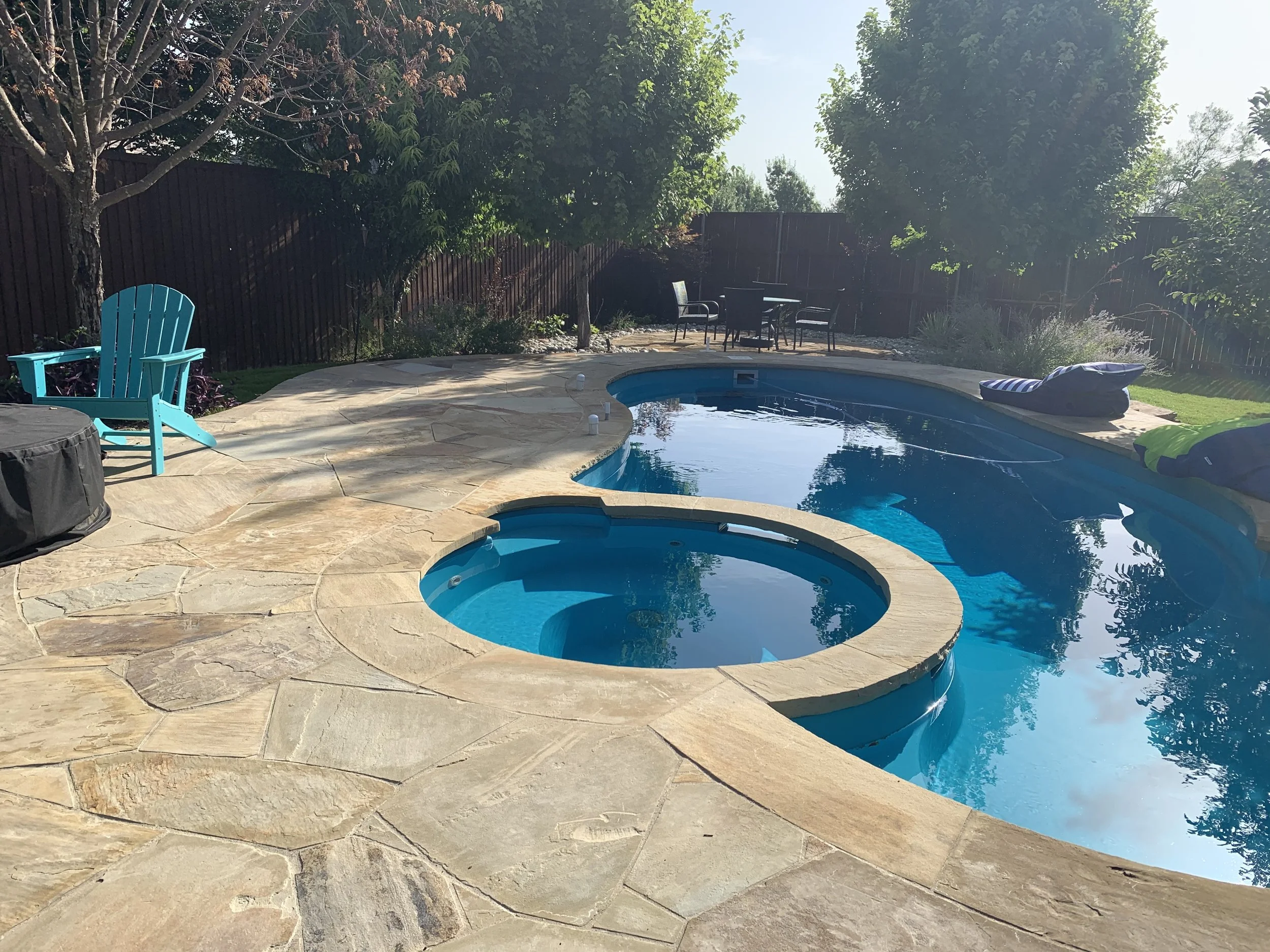 Backyard scene with a swimming pool and attached hot tub, surrounded by a stone patio. A turquoise Adirondack chair is on the left, and there are trees and a table with chairs in the background.
