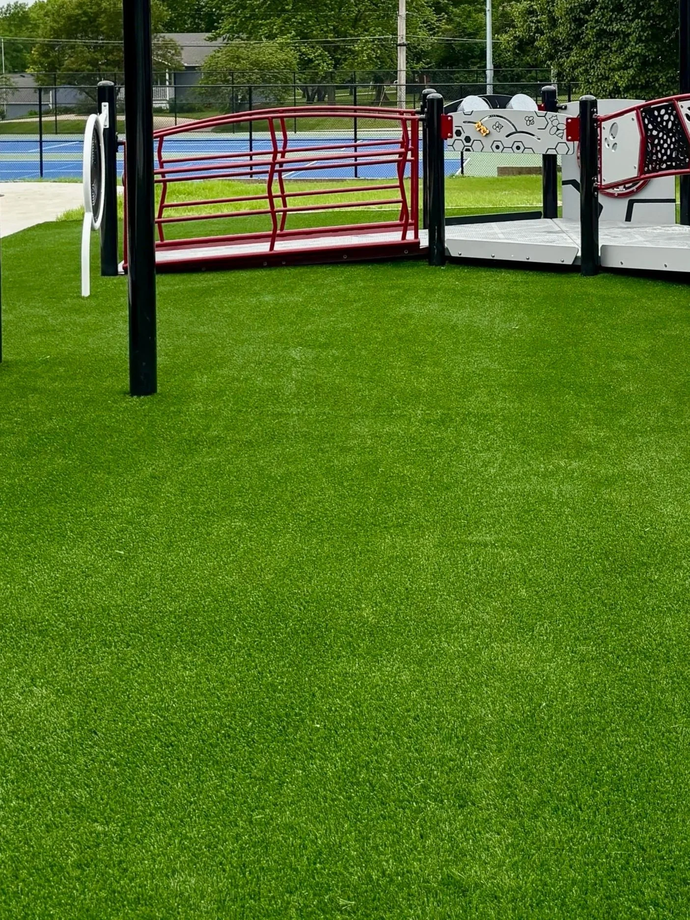 Modern playground with black and red climbing structures on artificial green turf, overcast sky, with trees and a sports court in the background.