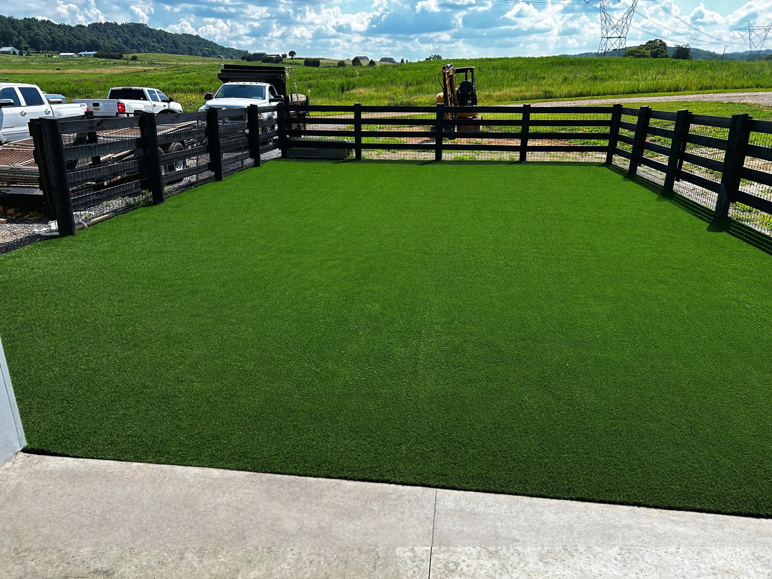 Artificial green grass yard with black wooden fence and surrounding construction equipment and vehicles, open field in the background under partly cloudy sky.
