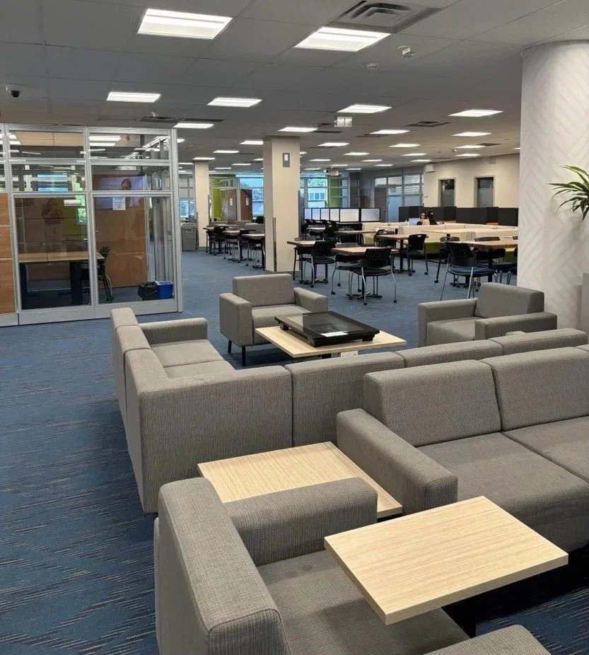 An empty modern office lounge area with gray sofas, wooden coffee tables, and a large open workspace with tables and chairs in the background.