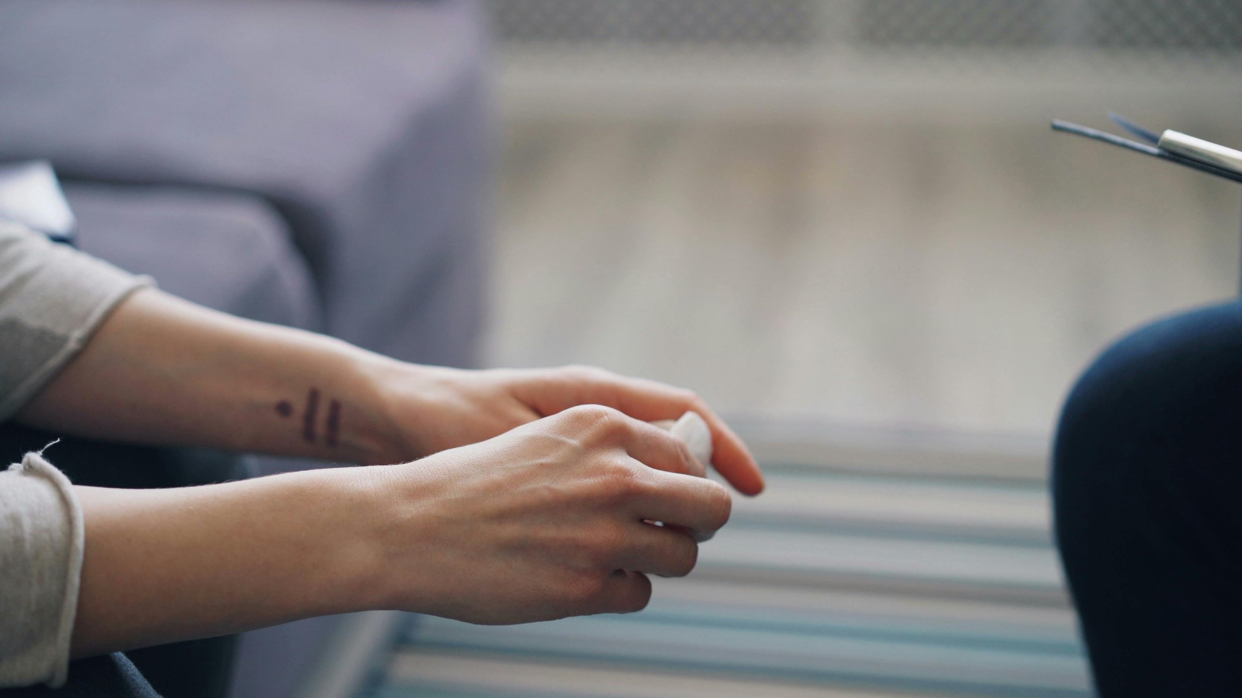 Close-up of a person's hands resting on their lap, with a tattoo visible on the wrist, and a blurred background.