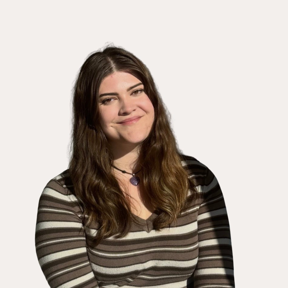 A young woman with long, wavy brown hair and fair skin, smiling softly, wearing a striped shirt and a purple heart necklace, set against a plain white background.