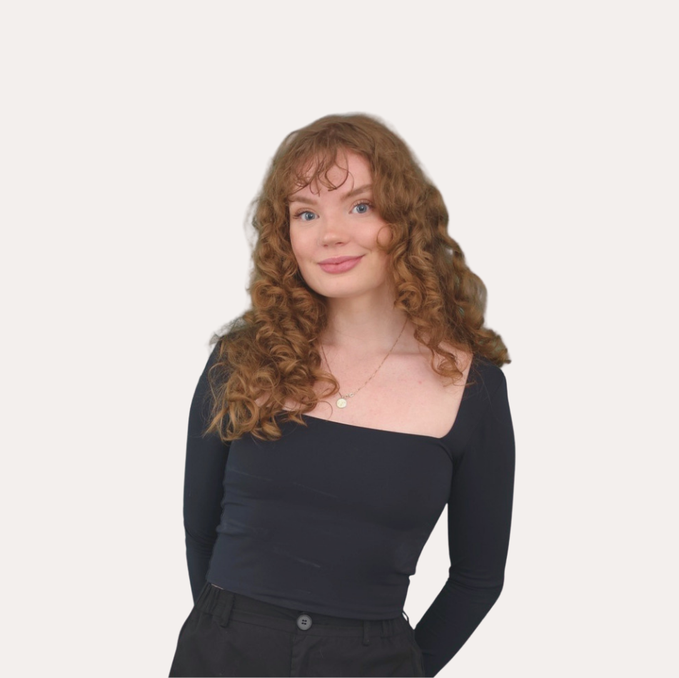 A woman with curly red hair wearing a black long sleeve top and a necklace, standing against a plain light background.