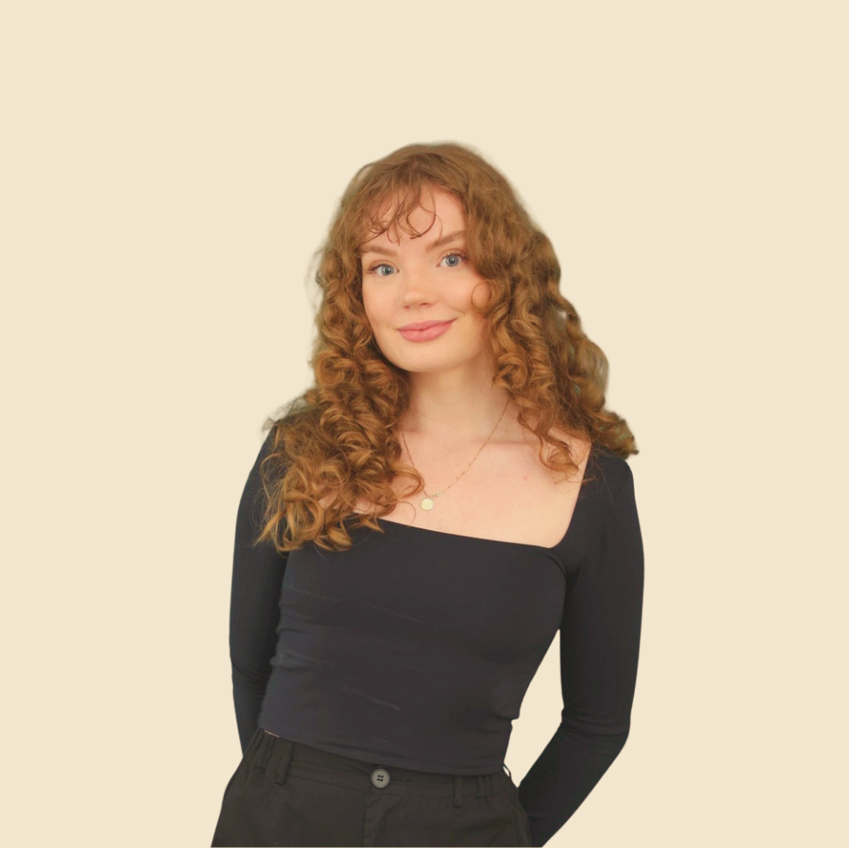 Young woman with long, curly red hair, wearing a black long-sleeve top and black pants, smiling softly, standing against a plain, light beige background.