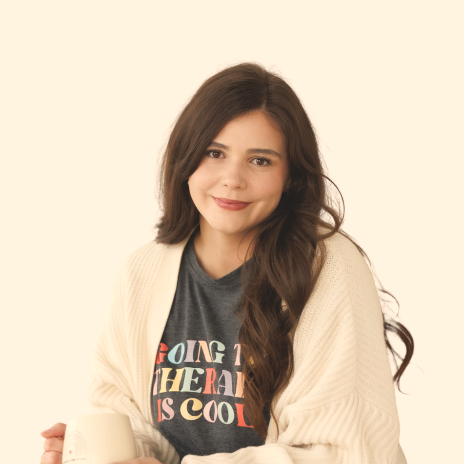 Young woman with long brown hair holding a white mug, wearing a gray T-shirt with colorful text and a cream cardigan, smiling against a light background.
