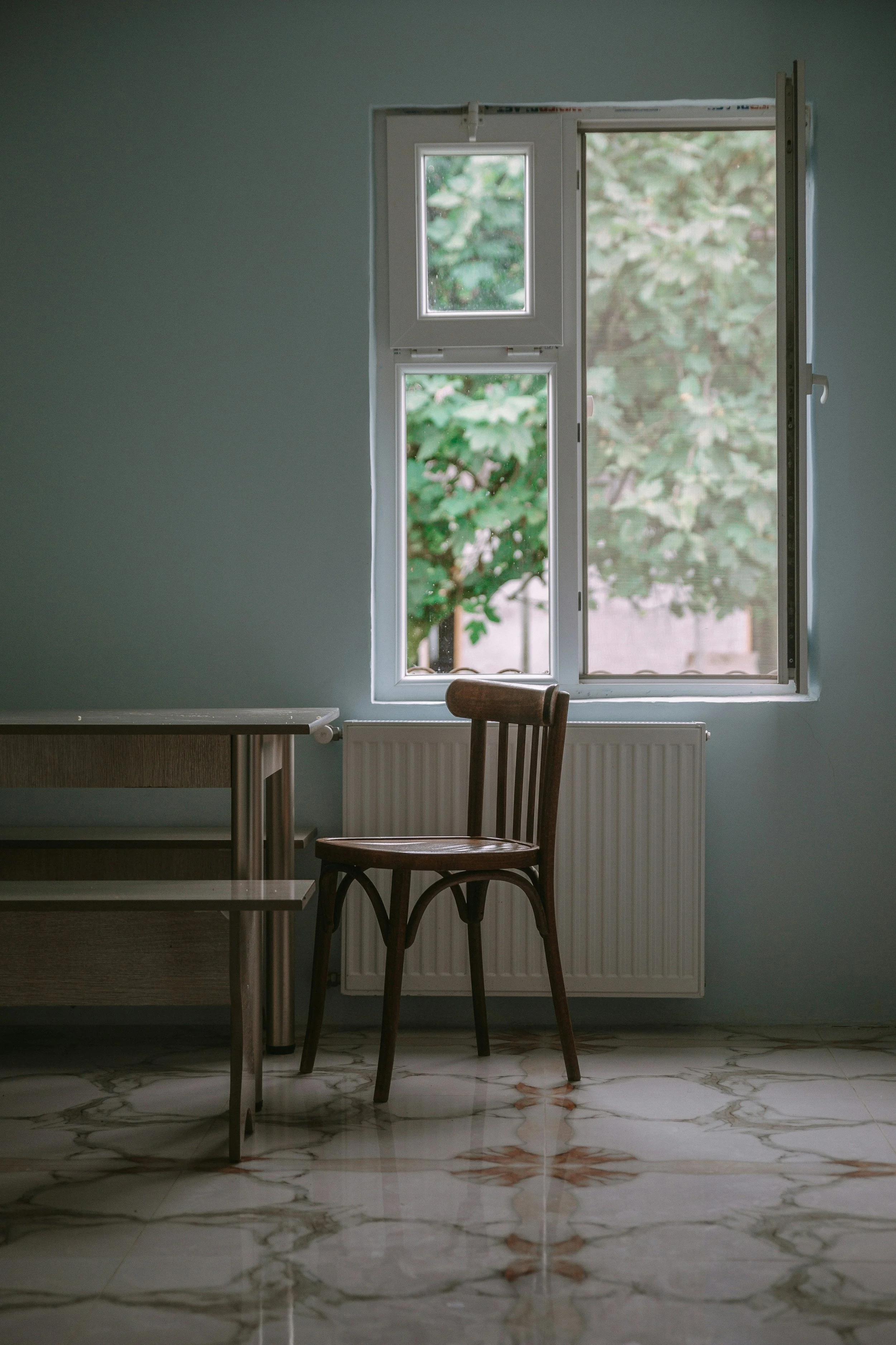 A window open to green trees, with a wooden chair in front of a radiator, a part of a kitchen countertop, and a marble floor.