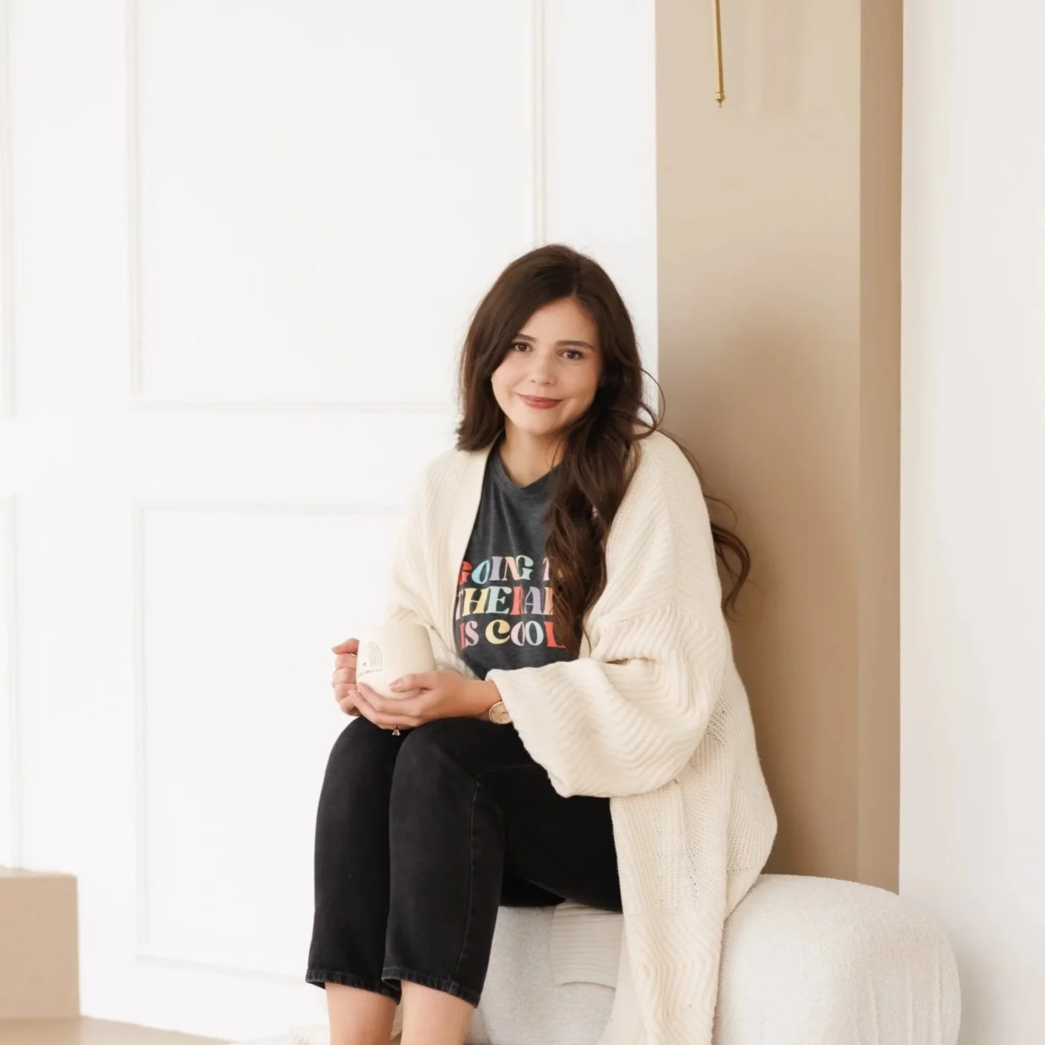 Young woman with long brown hair sitting on a white cushioned stool, smiling, wearing a gray graphic t-shirt, black pants, and a cream-colored cardigan, against a white wall background.