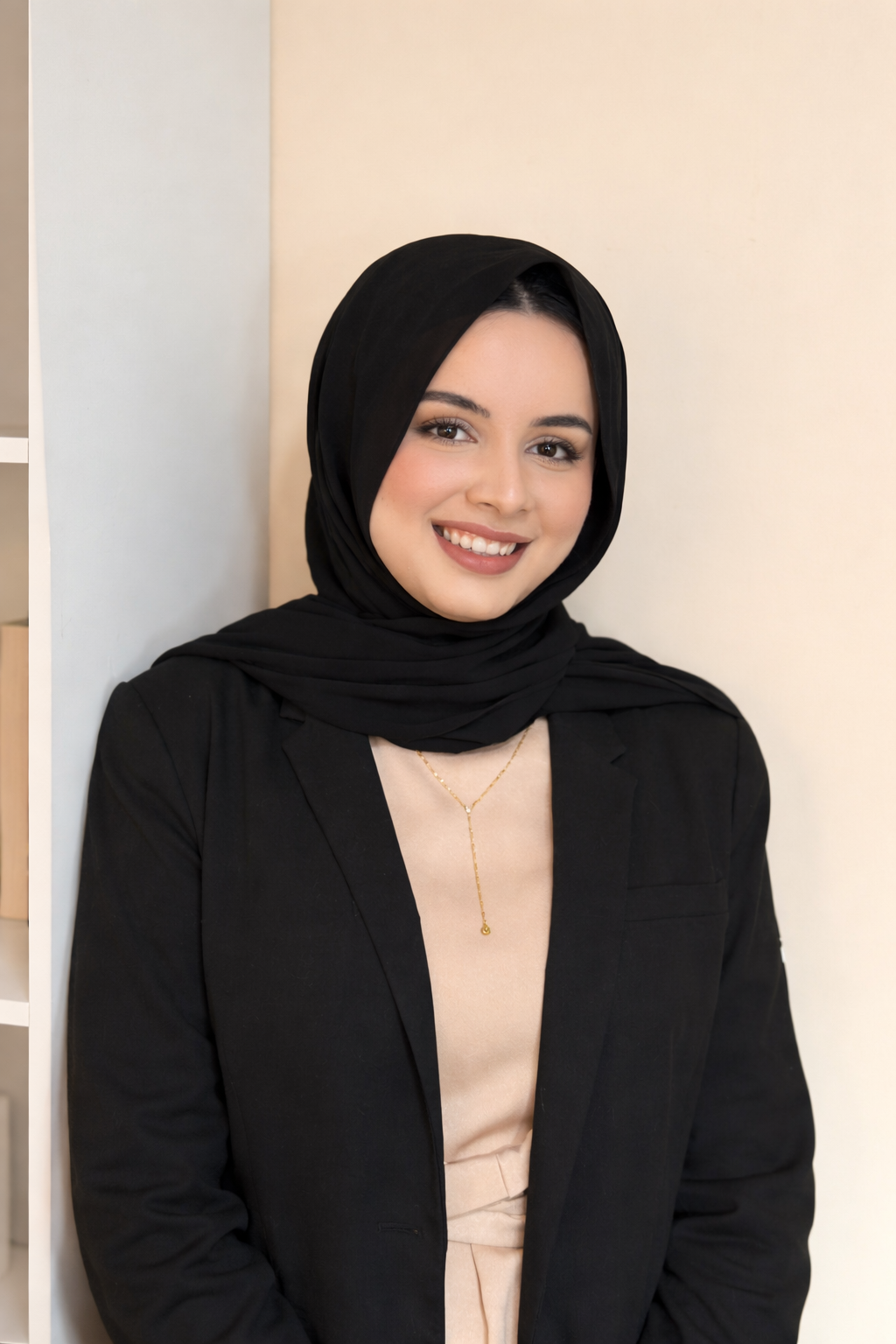 A young woman with light skin, smiling, wearing a black hijab, a black blazer, and a beige top, against a neutral background.