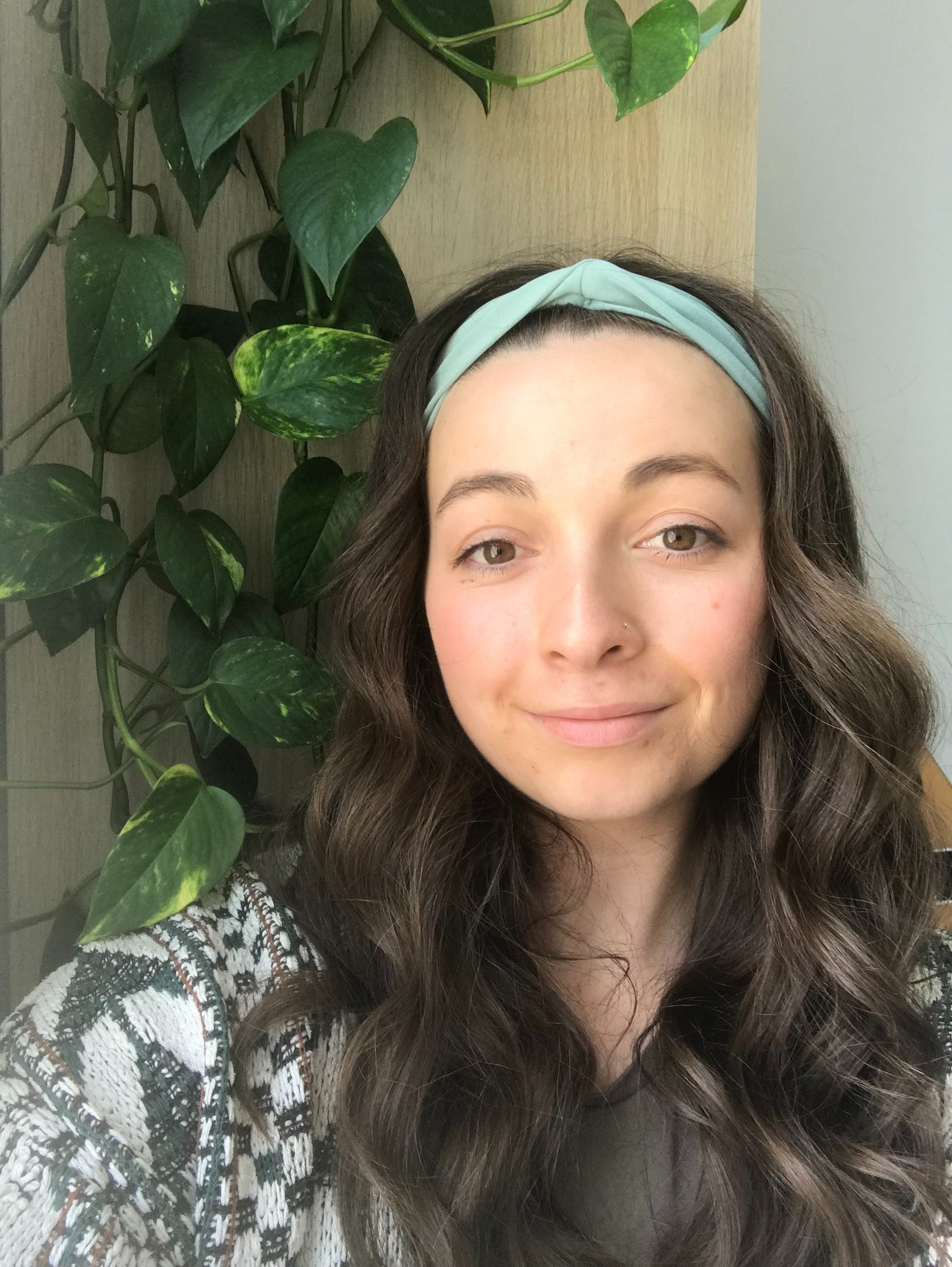 A woman with long wavy brown hair wearing a light green headband, a gray t-shirt, and a patterned cardigan, smiling at the camera against a plain, light-colored background.