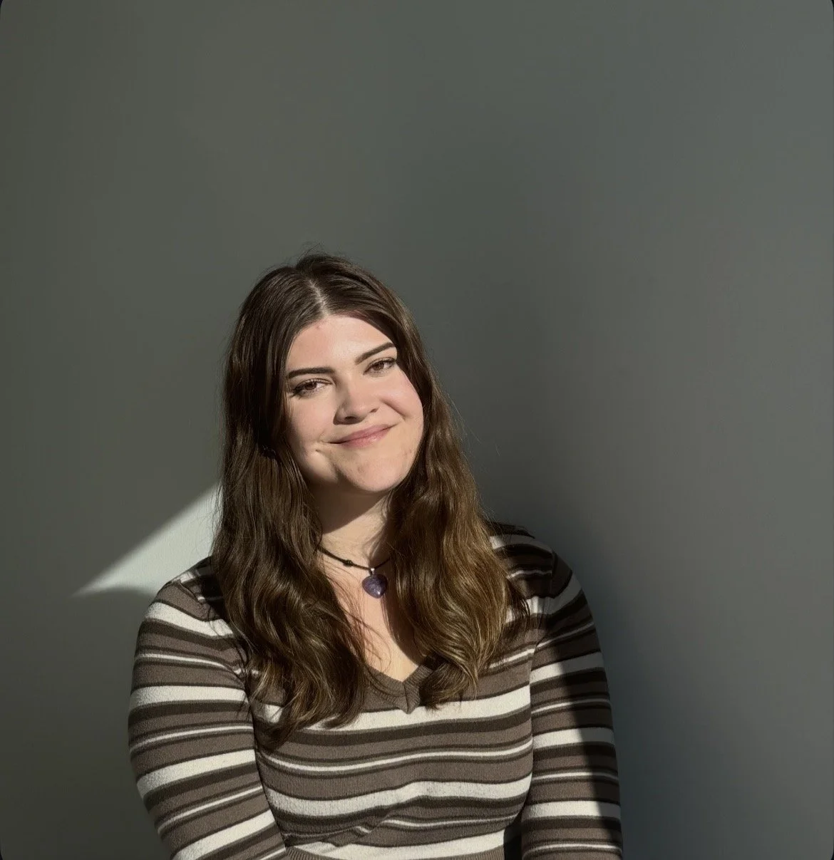 A young woman with long brown hair smiling, wearing a striped shirt and a purple heart-shaped necklace, against a plain light background.