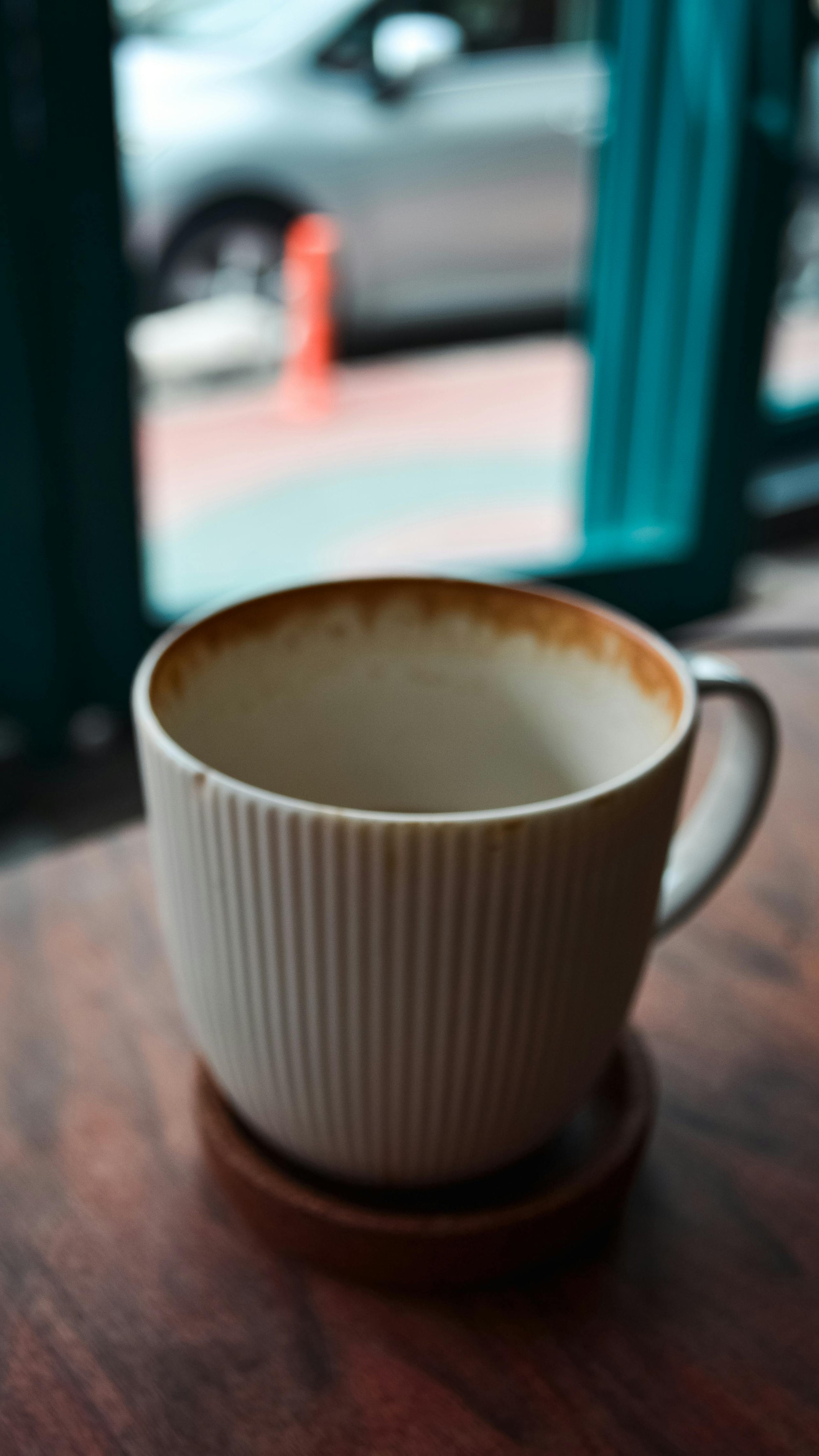 A ceramic mug with vertical ridges, filled with a beverage, sitting on a wooden coaster on a table. In the background, a window shows a blurred view of a car and an orange traffic cone outside.