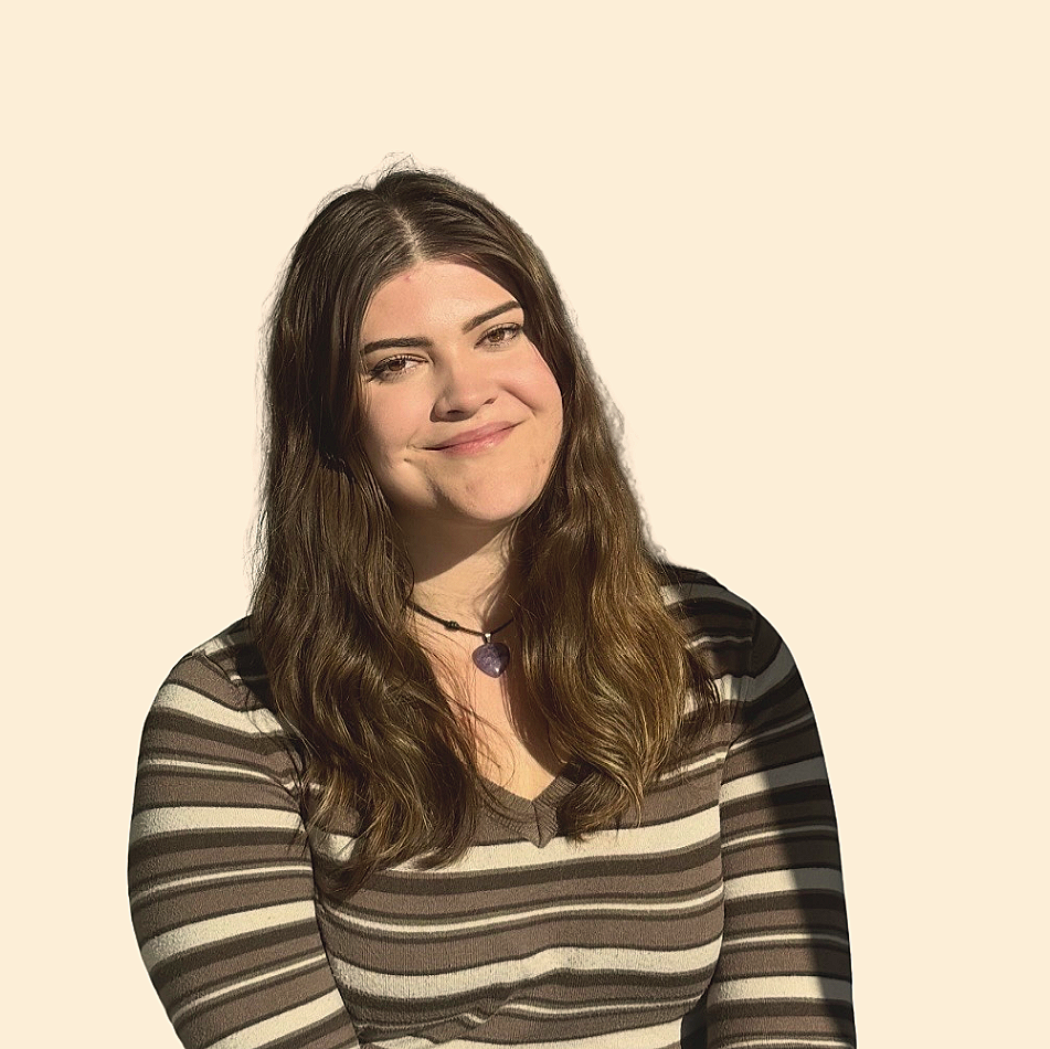 A young woman with long brown hair smiling, wearing a striped shirt and a purple heart-shaped necklace, against a plain light background.
