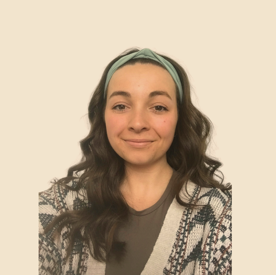 A woman with long wavy brown hair wearing a light green headband, a gray t-shirt, and a patterned cardigan, smiling at the camera against a plain, light-colored background.