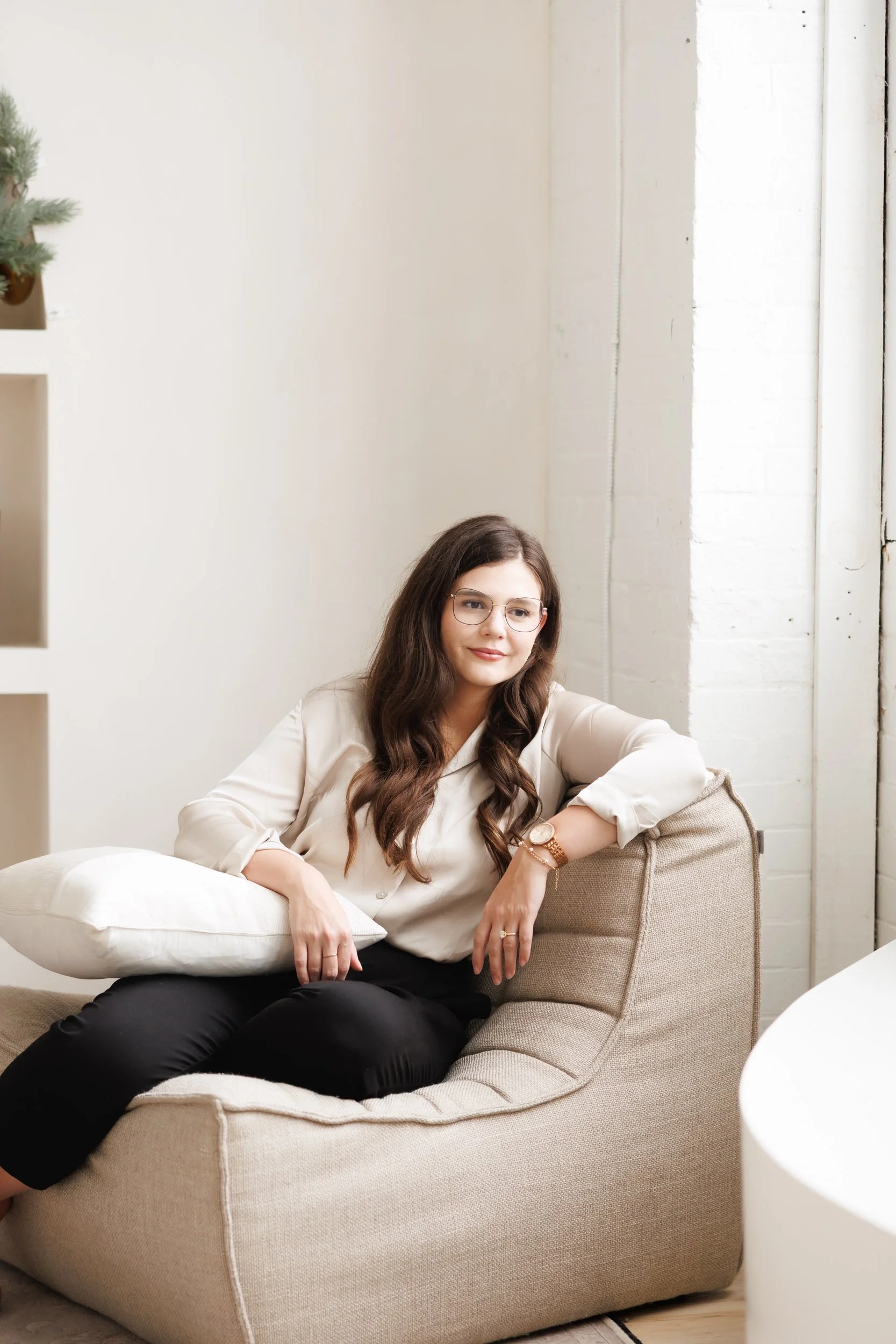 A woman with long brown hair, glasses, and a beige shirt sitting on a beige armchair, smiling slightly, in a bright room with white walls and a large window, with a white cushion on her lap.