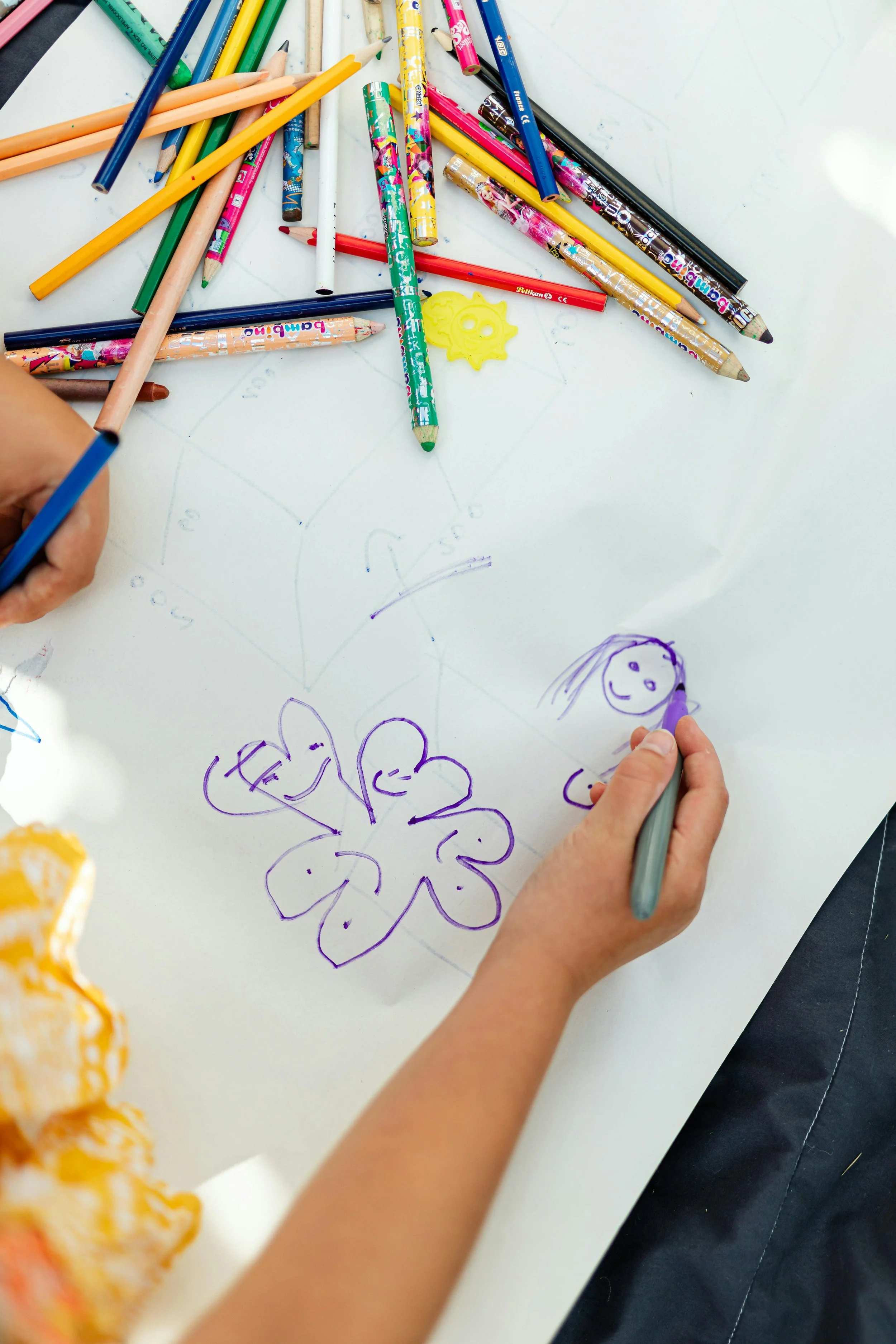 Hand drawing flowers and a smiling face with a purple marker on a white sheet of paper, surrounded by colored pencils and markers.