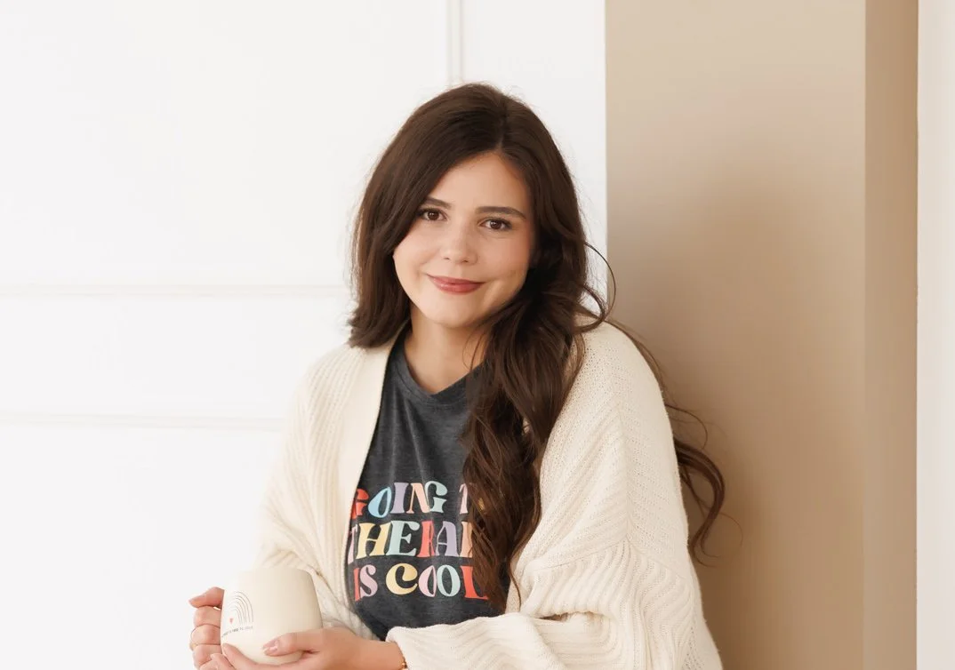 Young woman with long brown hair holding a white mug, wearing a gray T-shirt with colorful text and a cream cardigan, smiling against a light background.