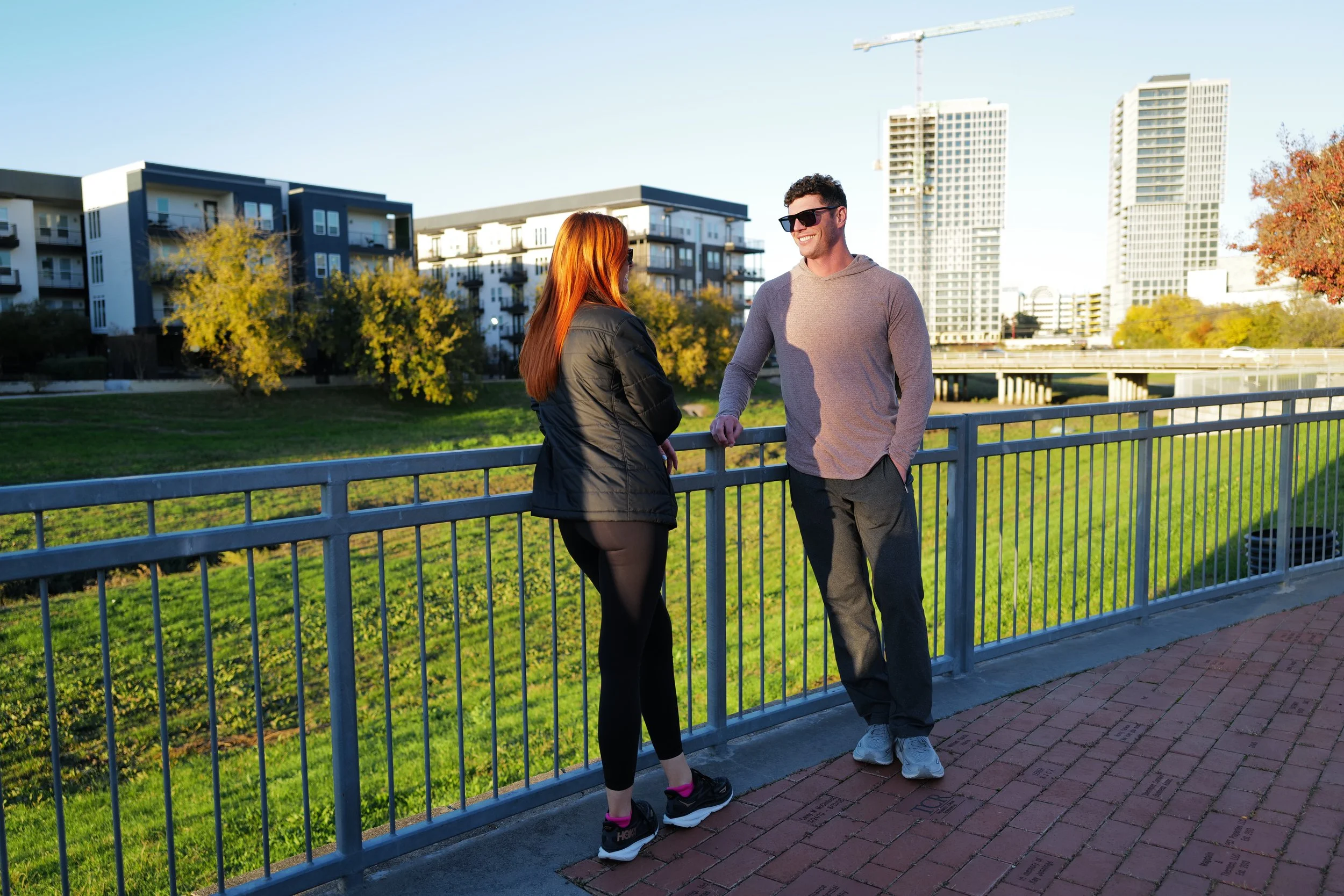 A man and woman talking near a metal railing on a brick pathway in a city park during late afternoon with tall buildings in the background.