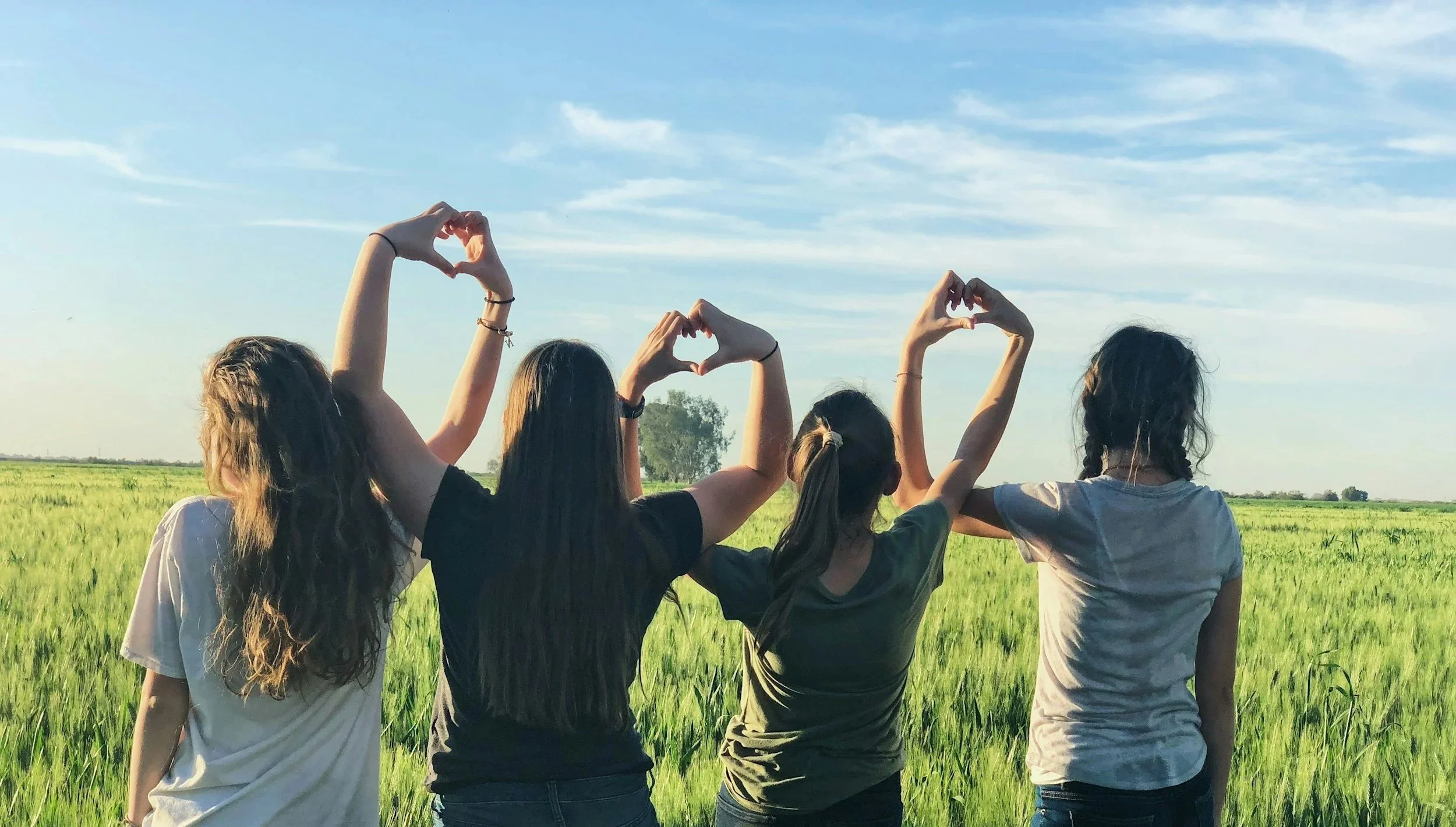 Four girls standing in a field with their backs facing the camera, making heart shapes with their hands against a blue sky with clouds.