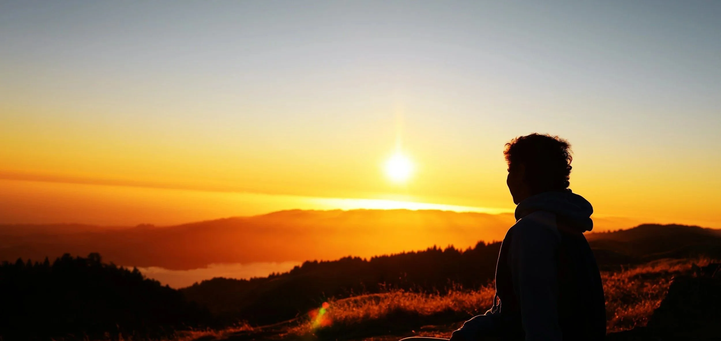 Person with curly hair sitting outdoors during sunset, silhouetted against a colorful sky with orange and yellow hues, overlooking rolling hills and mountains.