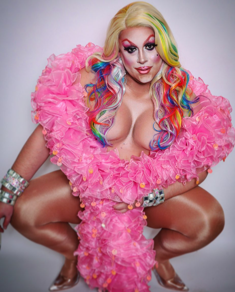 A person with long, rainbow-colored hair, wearing heavy makeup, in a pink ruffled dress with a deep neckline. They are squatting with one hand on their knee, against a plain white background.