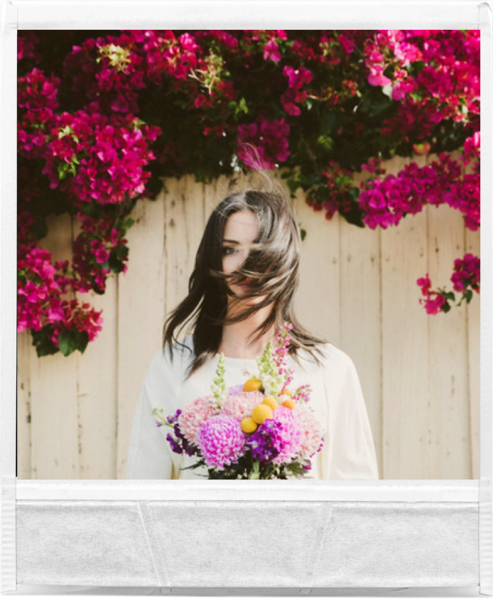 A woman holding a vibrant bouquet of flowers in front of a white fence, surrounded by lush pink bougainvillea