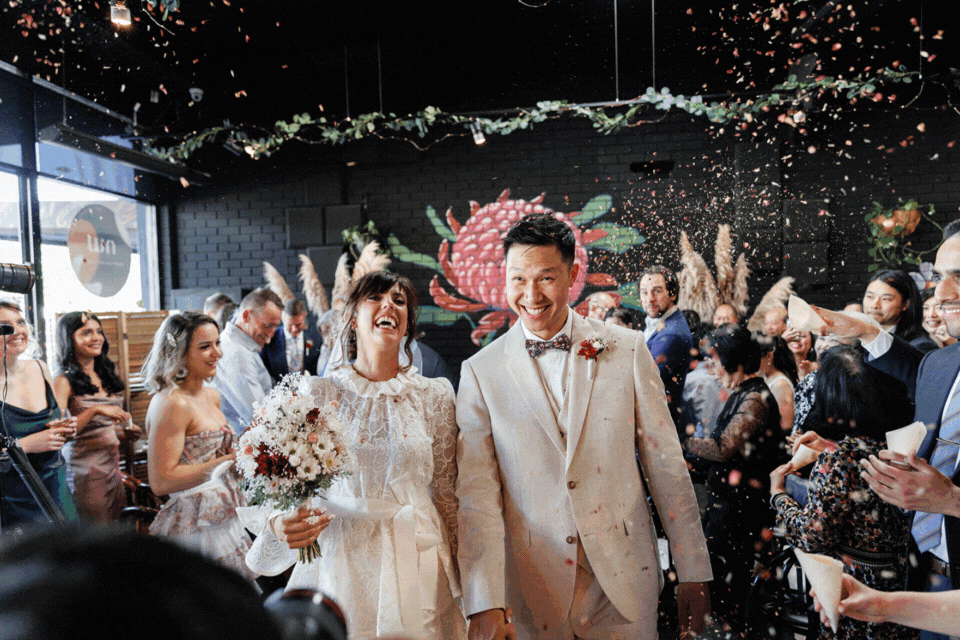 Bec and Nick walking through confetti at their Northside Wines wedding in Northcote, smiling as guests cheer them on.