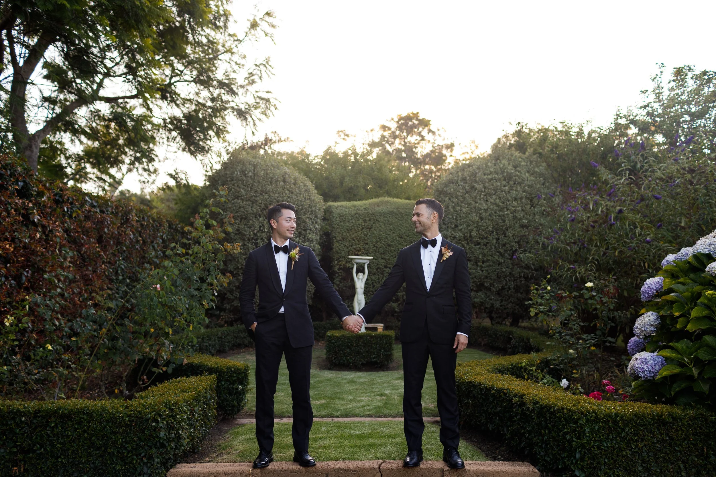 Oliver and Charlie holding hands at their Rivendell Winery Estate wedding in Yallingup, dressed in matching black tuxedos.