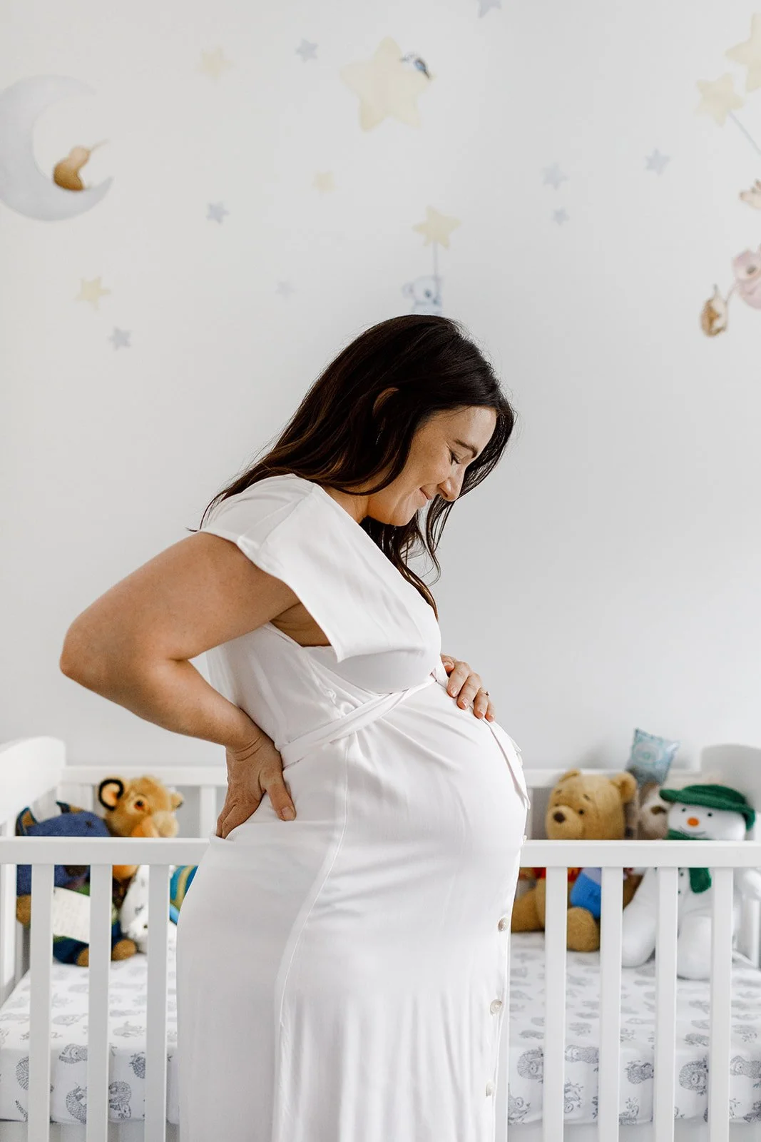 Meg gently cradling her pregnant belly while standing in the nursery, smiling in anticipation.