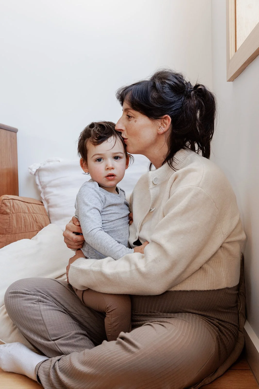 Greta sitting with her baby at home, gently holding and kissing them on the head in a quiet, tender moment.