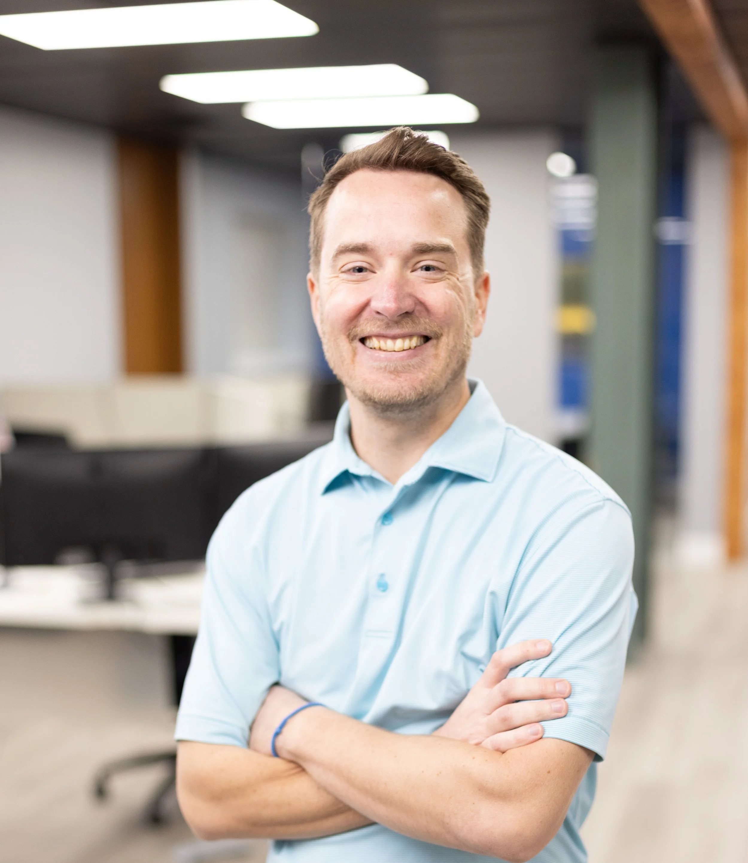 A smiling man with arms crossed, wearing a light blue polo shirt, standing in an office environment with computer desks and chairs in the background.