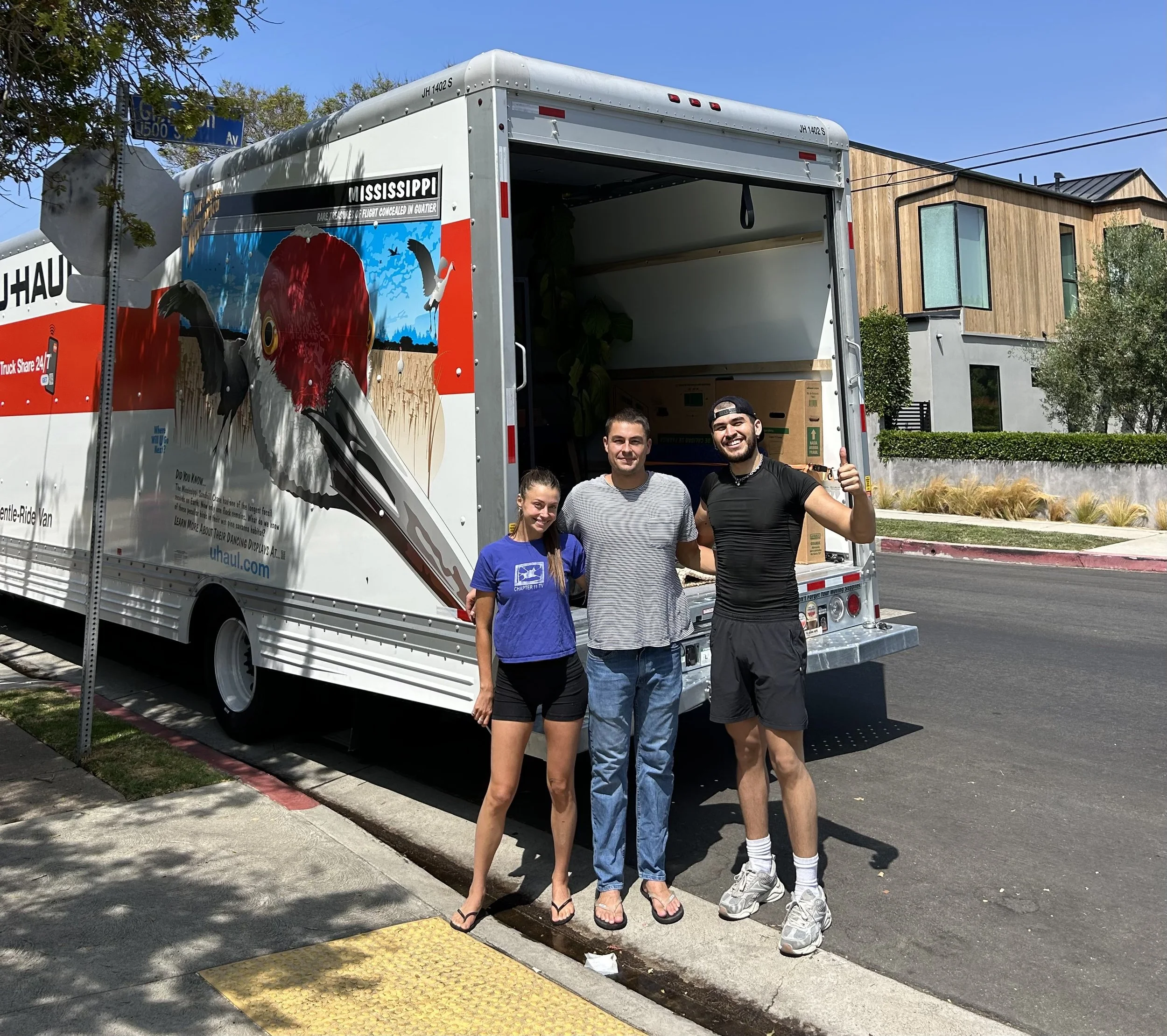 Three people standing in front of a U-Haul truck, smiling. The truck has a Mississippi bird graphic on its side. The group appears to be moving or moving in, with boxes visible inside the truck.