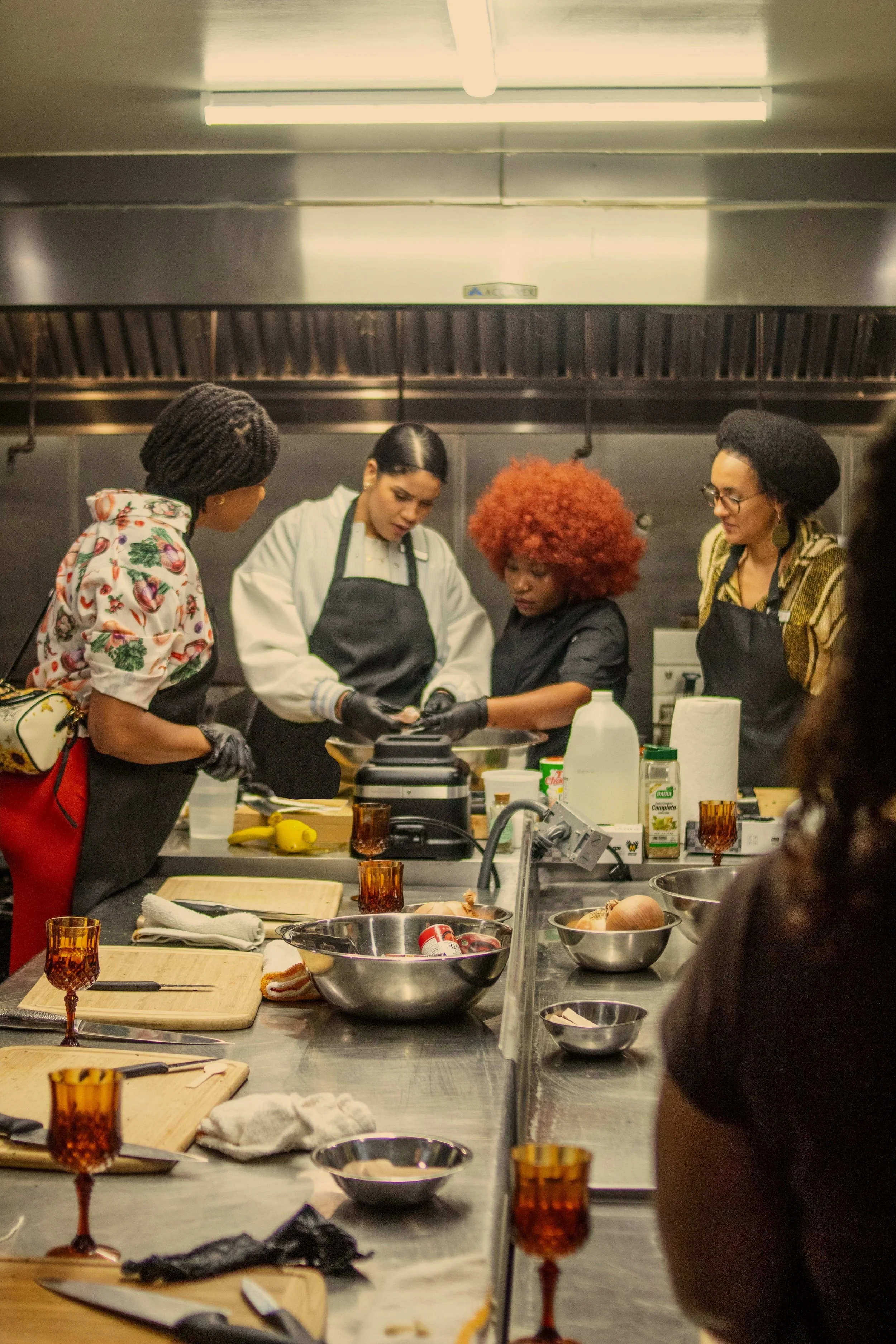 Four women in a professional kitchen preparing food during a Haitian cooking class, with ingredients and kitchen tools on the counter.