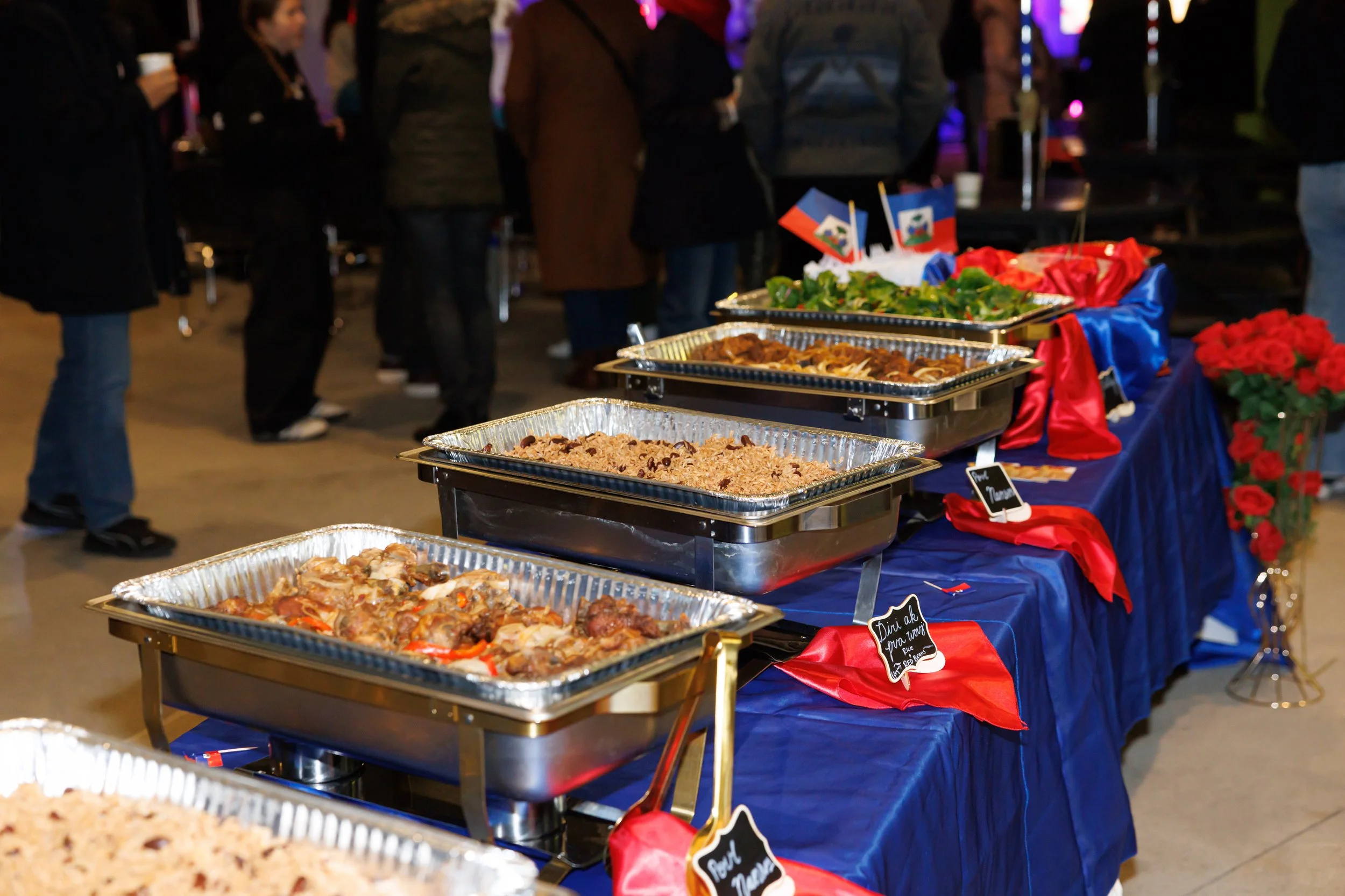 A group of people gathered around a table with charcuterie and snacks, engaging in conversation at a social event. The table has assorted fruits, crackers, and meats.