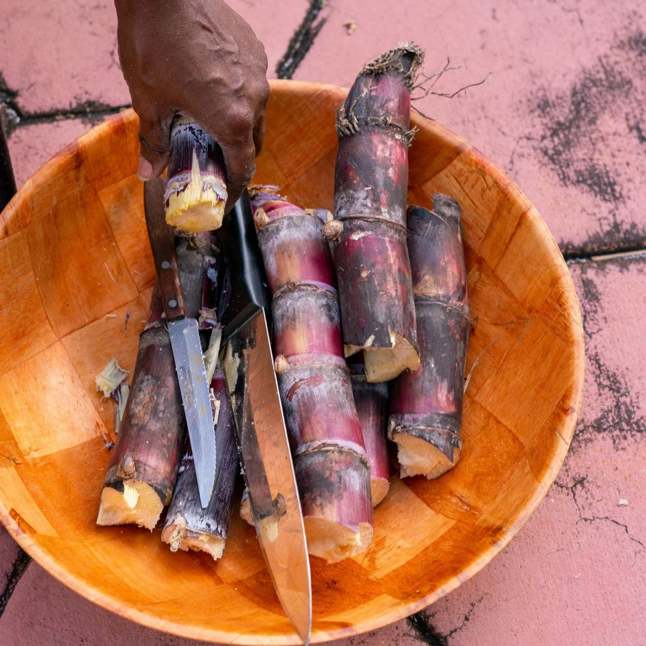 A hand holding a piece of sugarcane in a wooden bowl with more sugarcane pieces and two knives on a surface.
