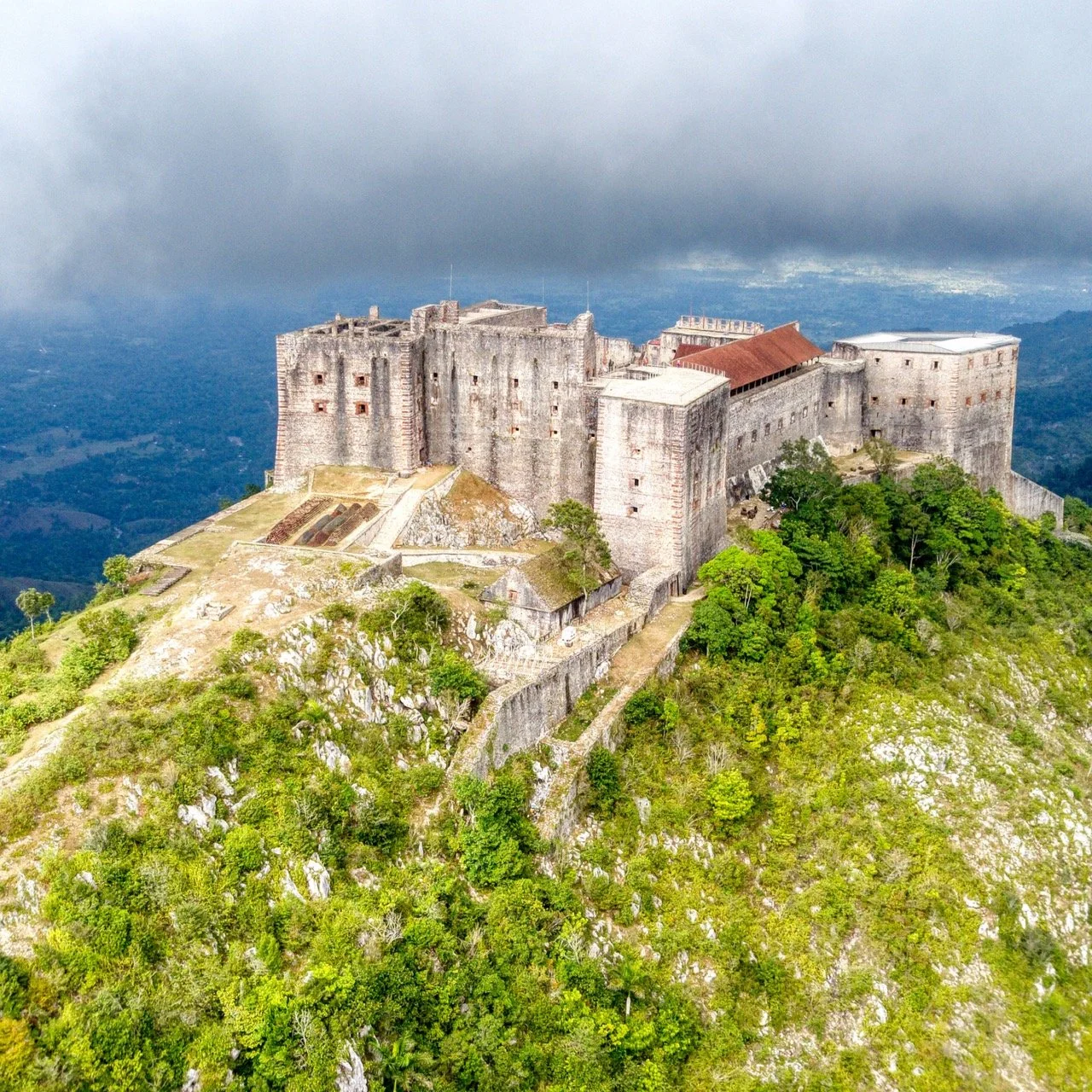 Aerial view of a large stone fortress on a hilltop surrounded by lush green landscape and cloudy sky.