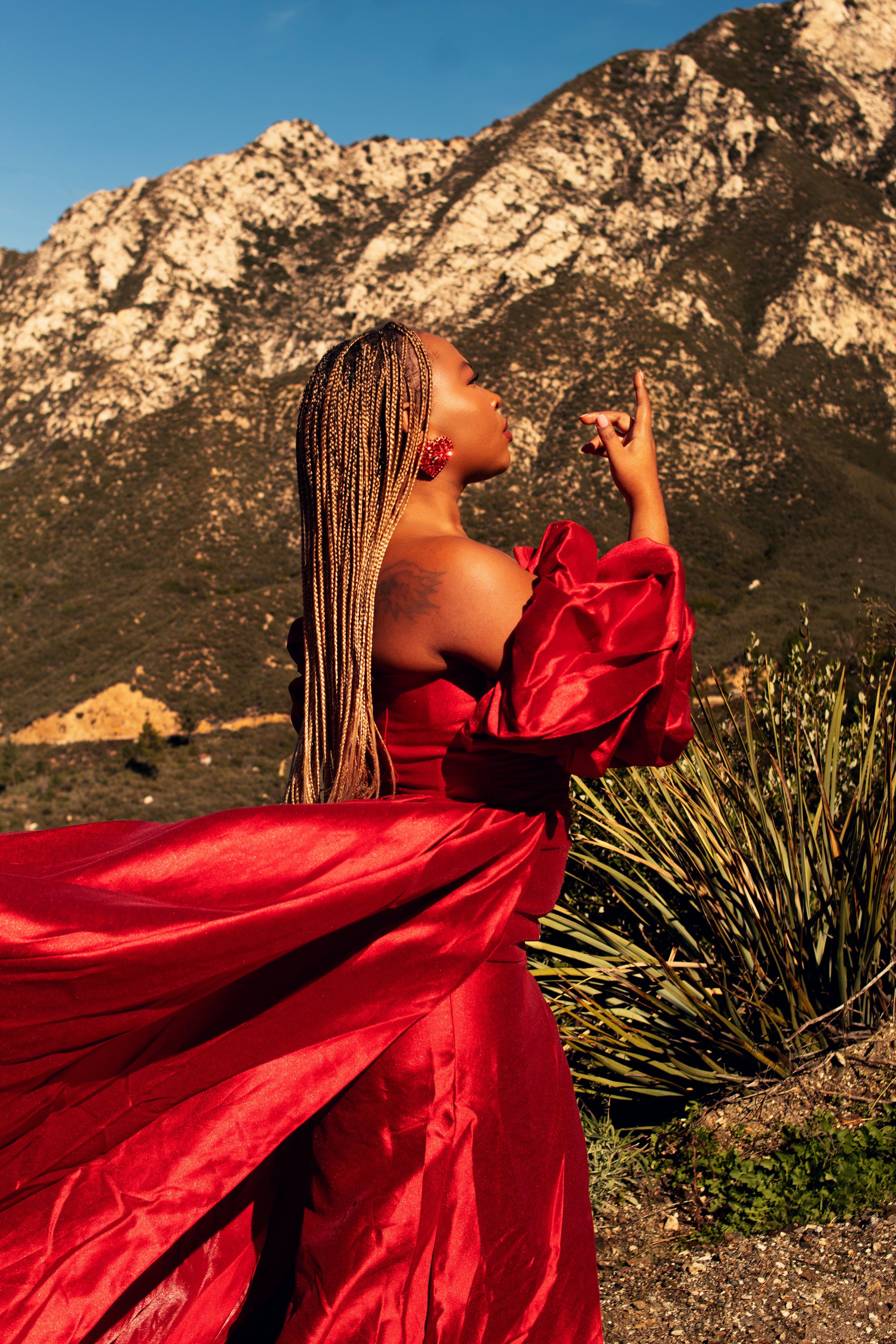 Kerline Ordeus in a flowing red gown standing outdoors with mountains in the background, touching her face with her eyes closed.