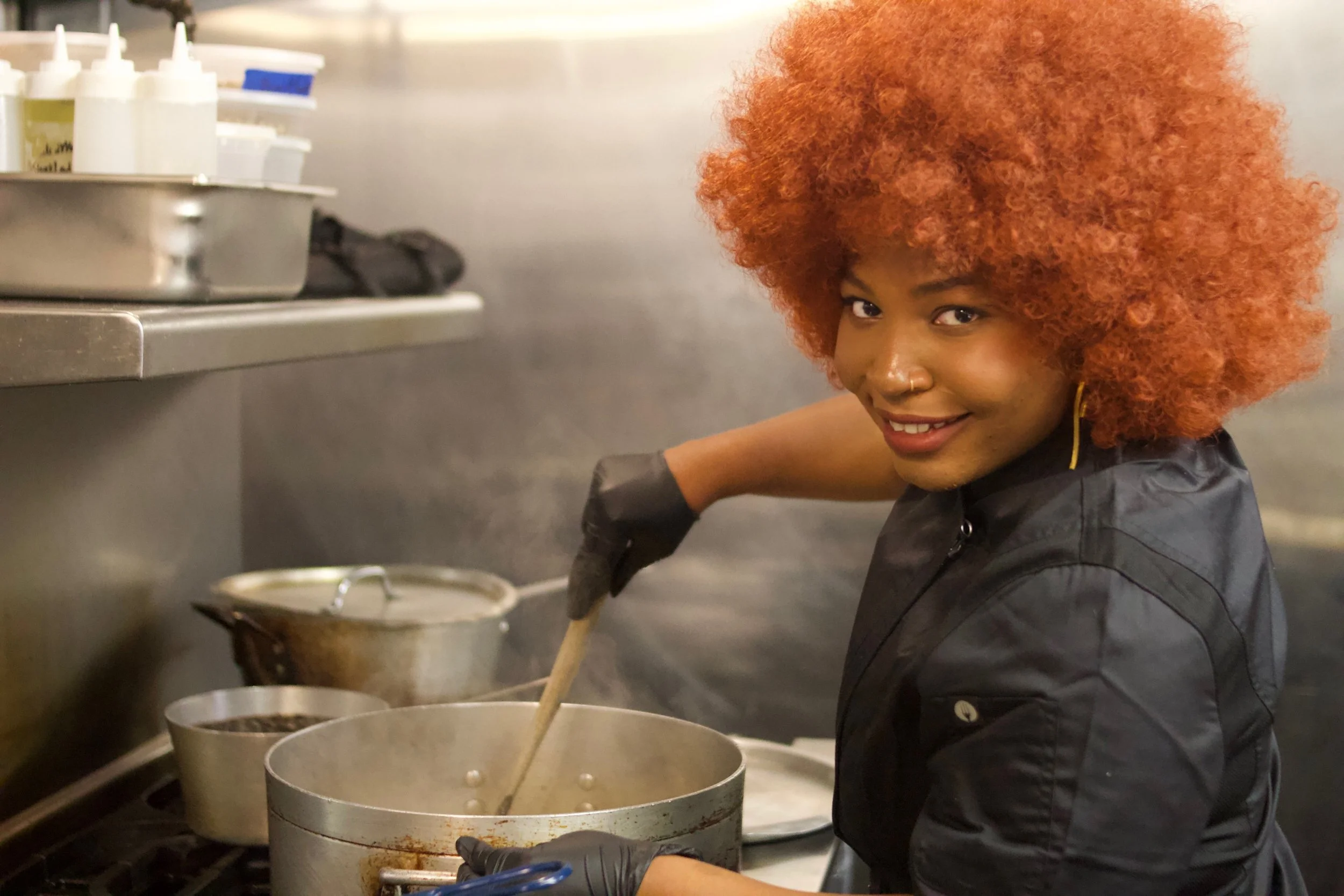 Kerline Ordeus with curly red hair cooking in a commercial kitchen, stirring a large pot.