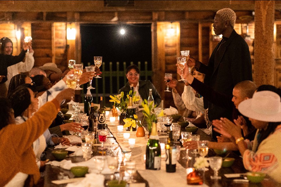 People raising glasses in toast around a long dining table during a celebration in a cozy, wood-paneled setting.