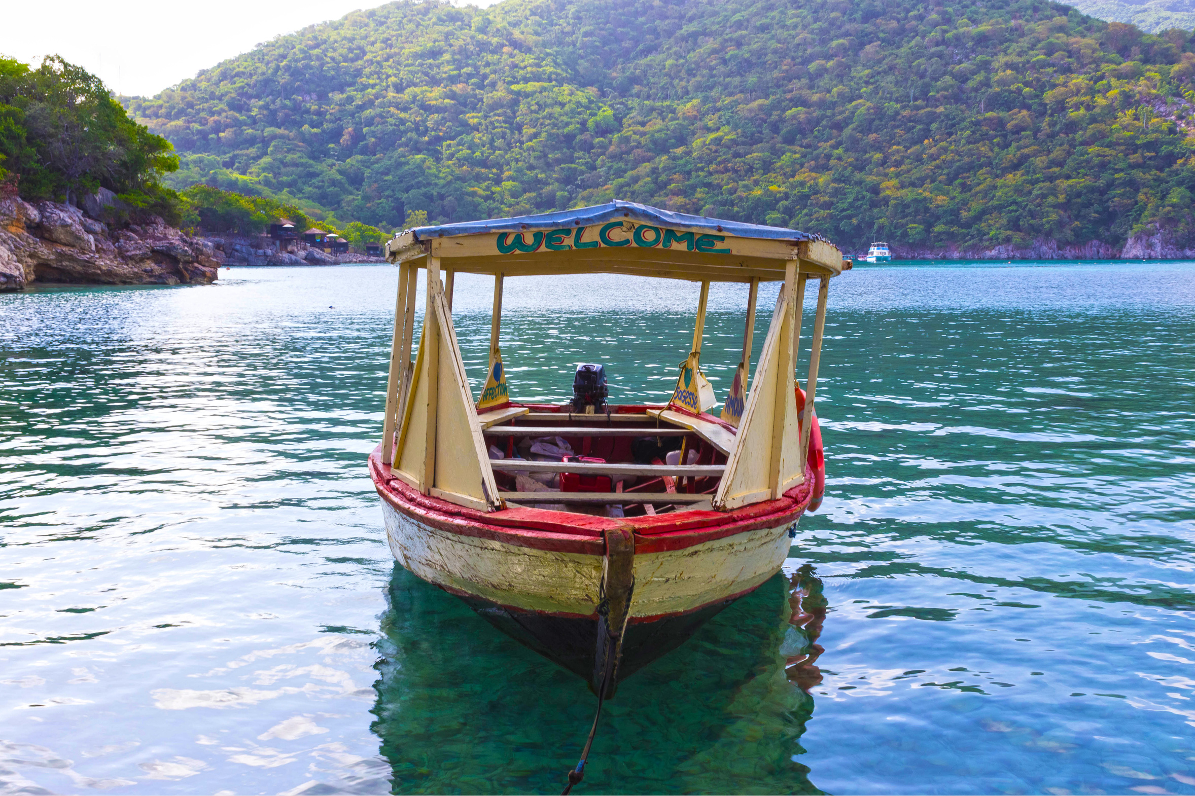 A wooden boat with a "Welcome" sign docked on clear turquoise water, surrounded by lush green hills and rocky shoreline.