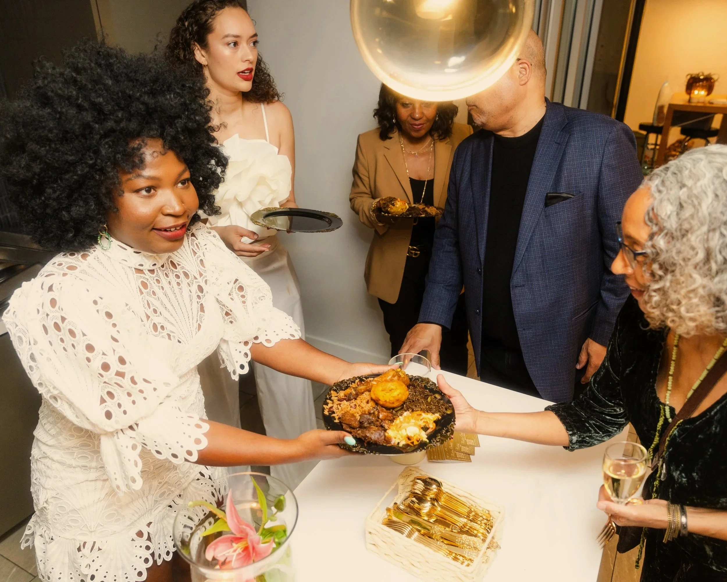 Group of diverse people at a celebration, with woman in white dress passing a plate of food to another woman holding a glass of wine.