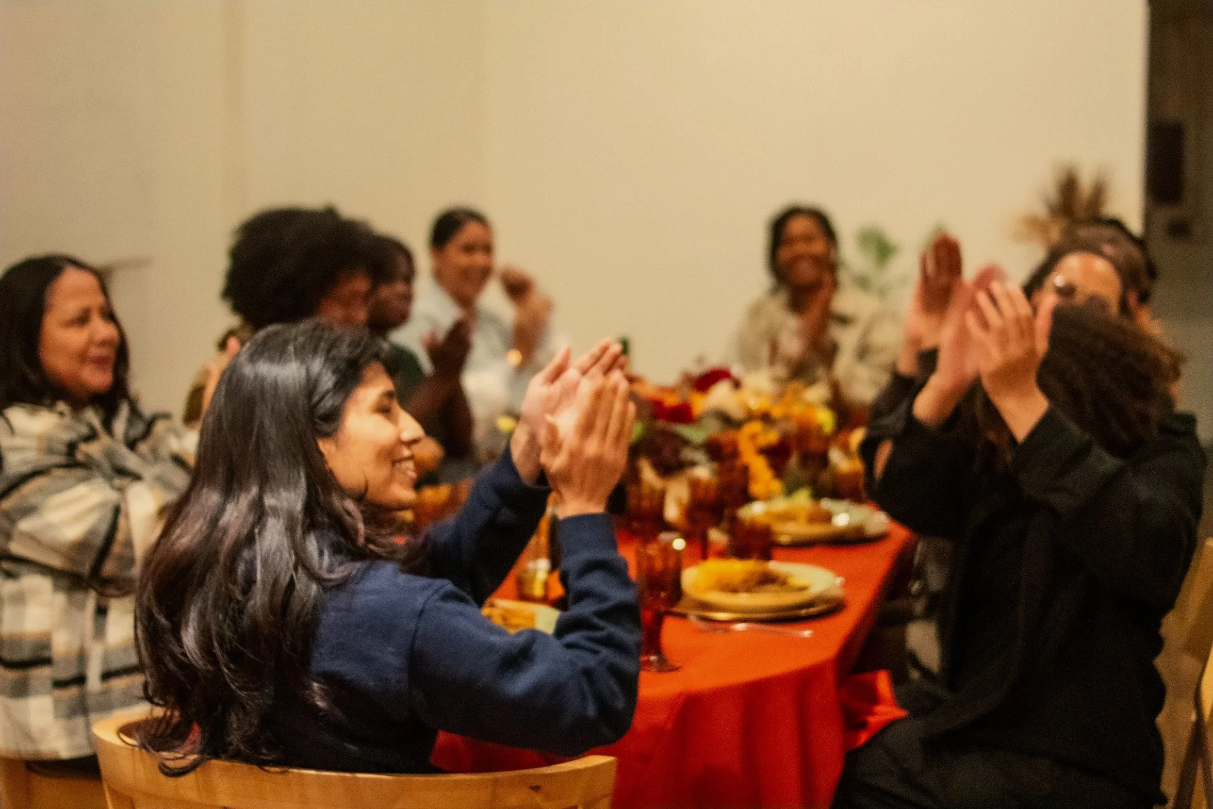 A group of people gathered around a table with charcuterie and snacks, engaging in conversation at a social event. The table has assorted fruits, crackers, and meats.