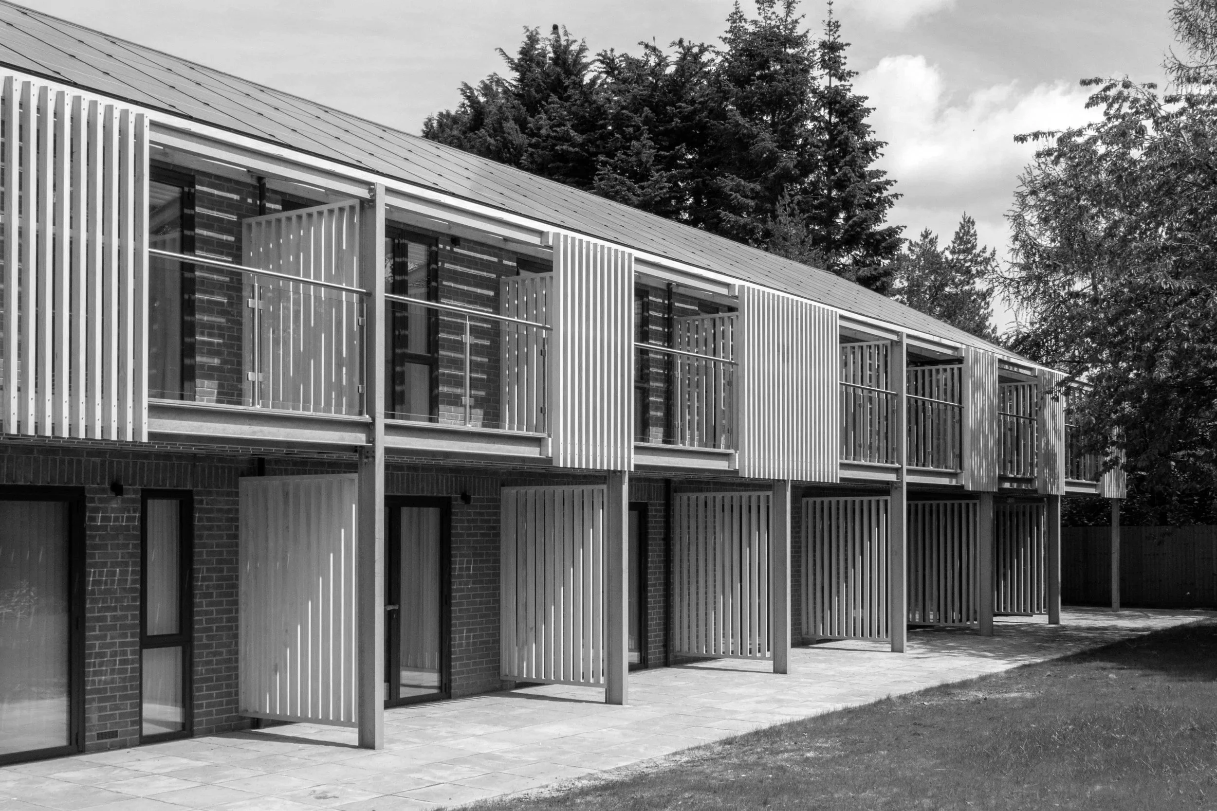Modern apartment building with brick walls, floor to ceiling glazing, and timber slatted privacy panels on balconies, surrounded by trees.